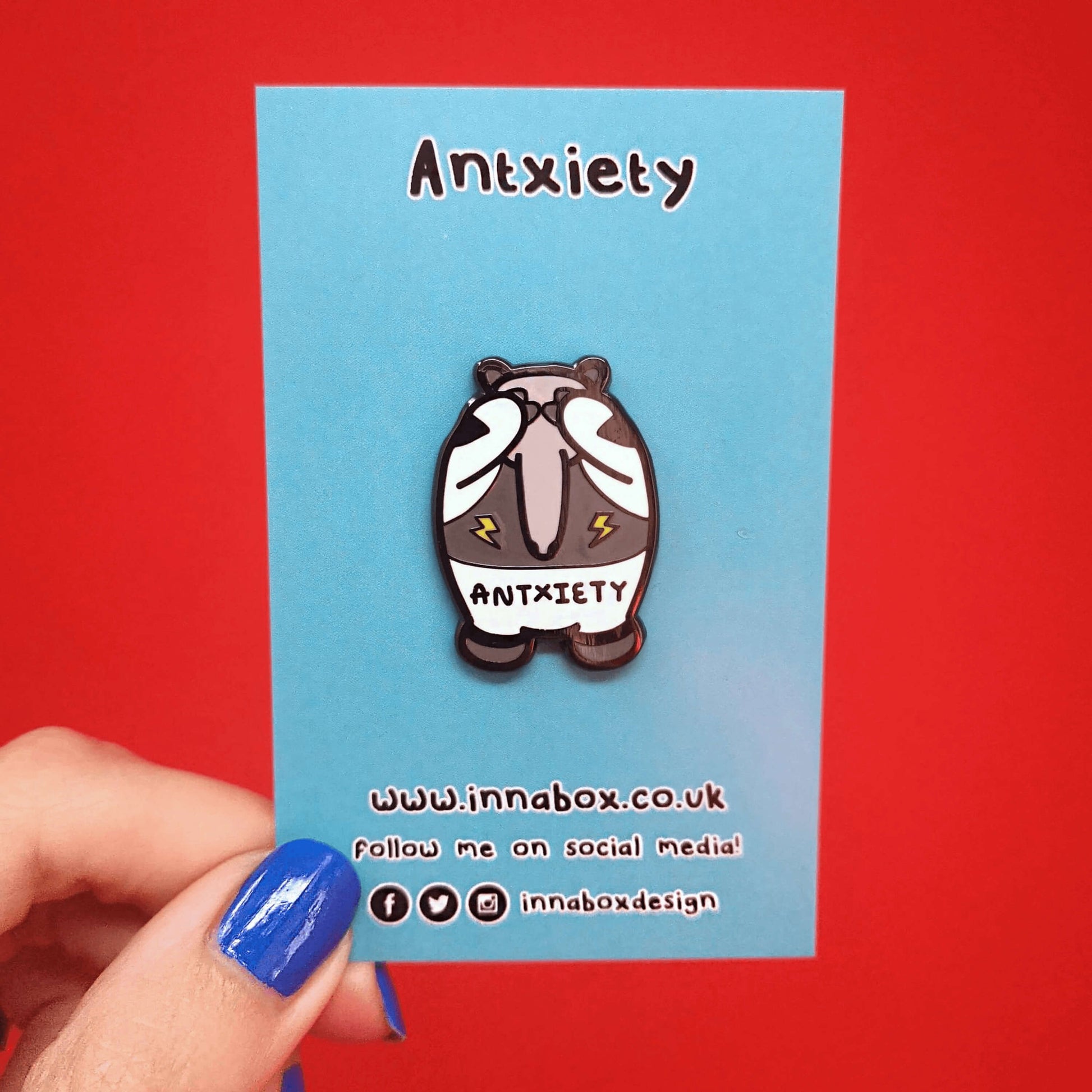 Antxiety Enamel Pin on a blue backing card being held up by a hand with blue nail varnish in front of a red background. The pin is a grey and white anteater with yellow lightning bolts and the word antxiety across its belly. The pin is designed to raise awareness for anxiety disorders and anxious emotions.