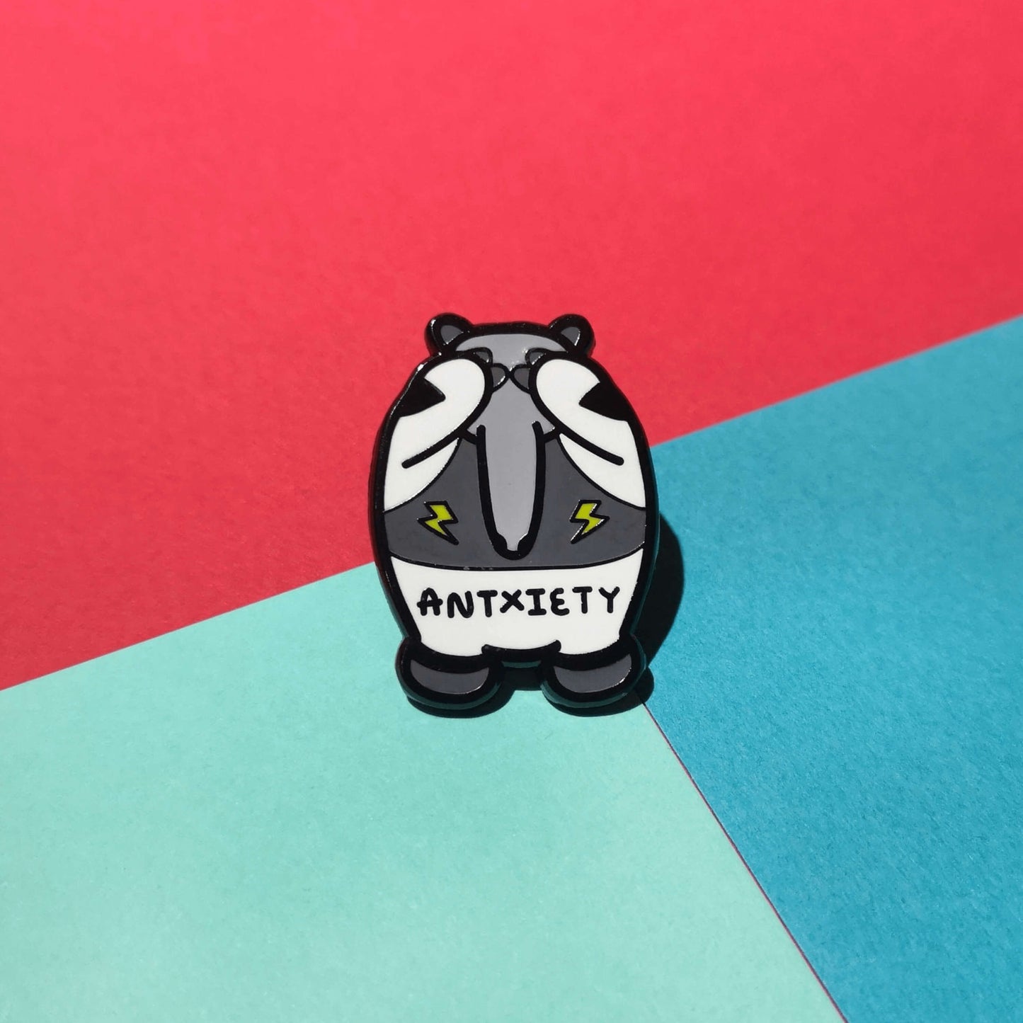 Antxiety Enamel Pin - Anxiety sat on a blue, green and red background. The pin is a grey and white anteater with yellow lightning bolts and the word antxiety across its belly. The pin is designed to raise awareness for anxiety disorders and anxious emotions.
