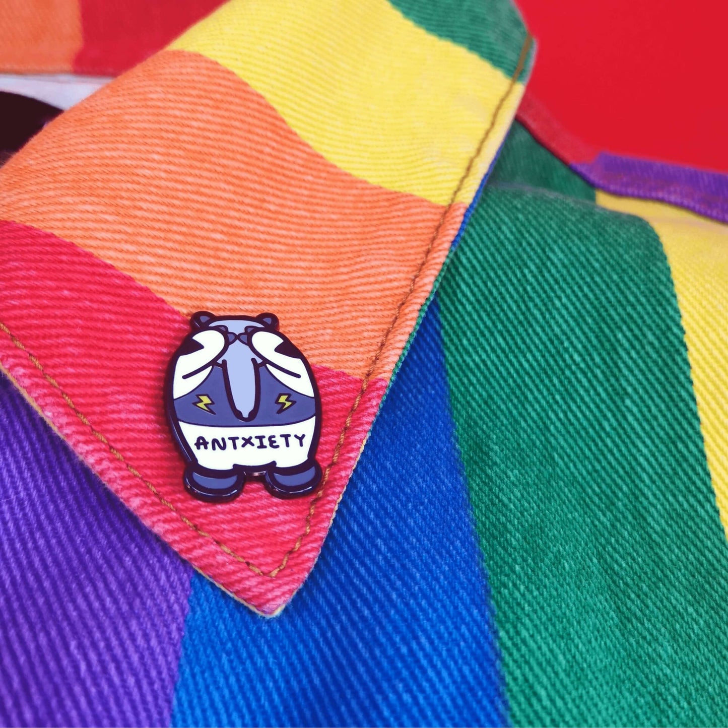 Antxiety Enamel Pin - Anxiety pinned on a rainbow jacket. The pin is a grey and white anteater with yellow lightning bolts and the word antxiety across its belly. The pin is designed to raise awareness for anxiety disorders and anxious emotions.