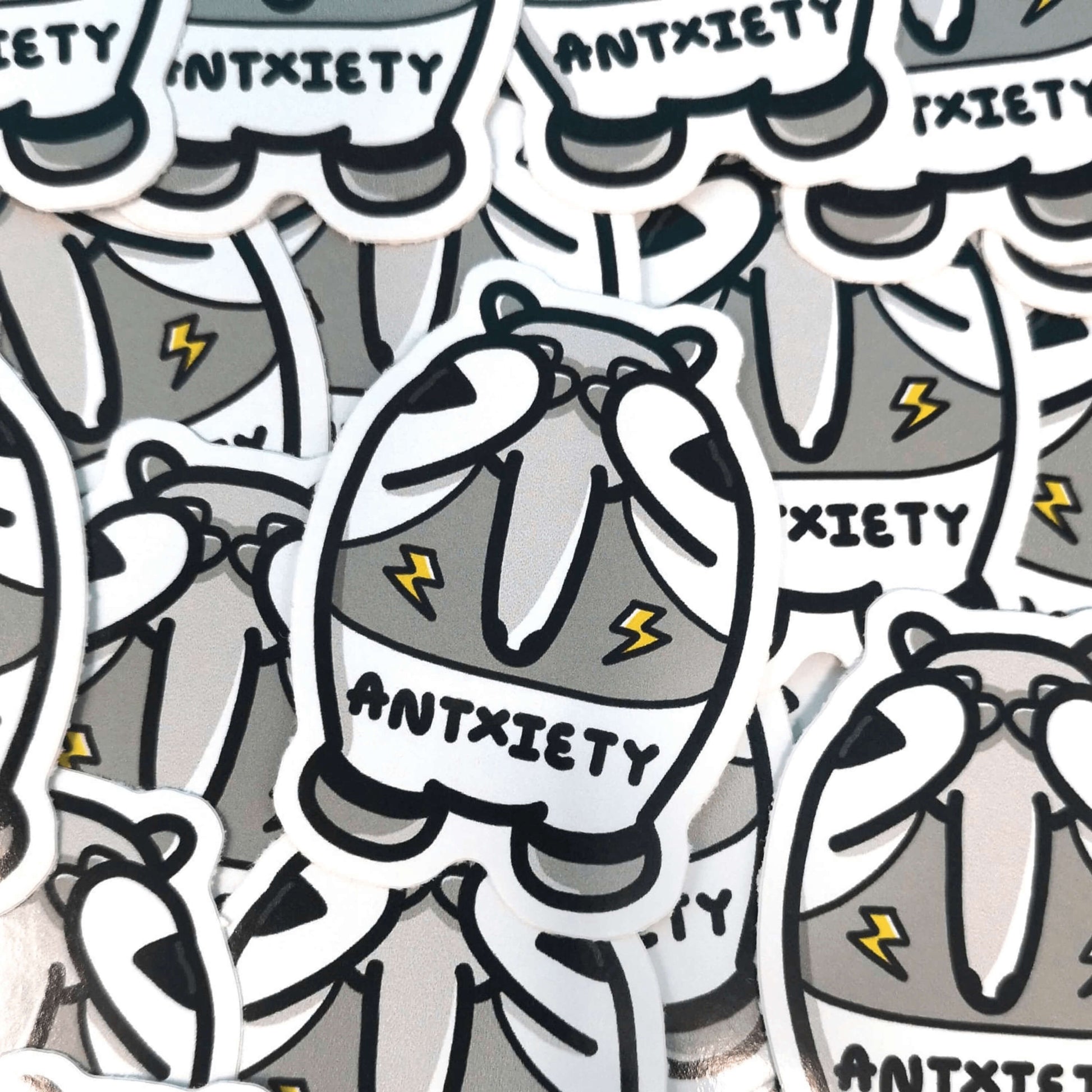 Antxiety Sticker - Anxiety sat on lots of the stickers. The sticker is a grey and white anteater with yellow lightning bolts and the word antxiety across its belly. The sticker is designed to raise awareness for anxiety disorders and anxious emotions.