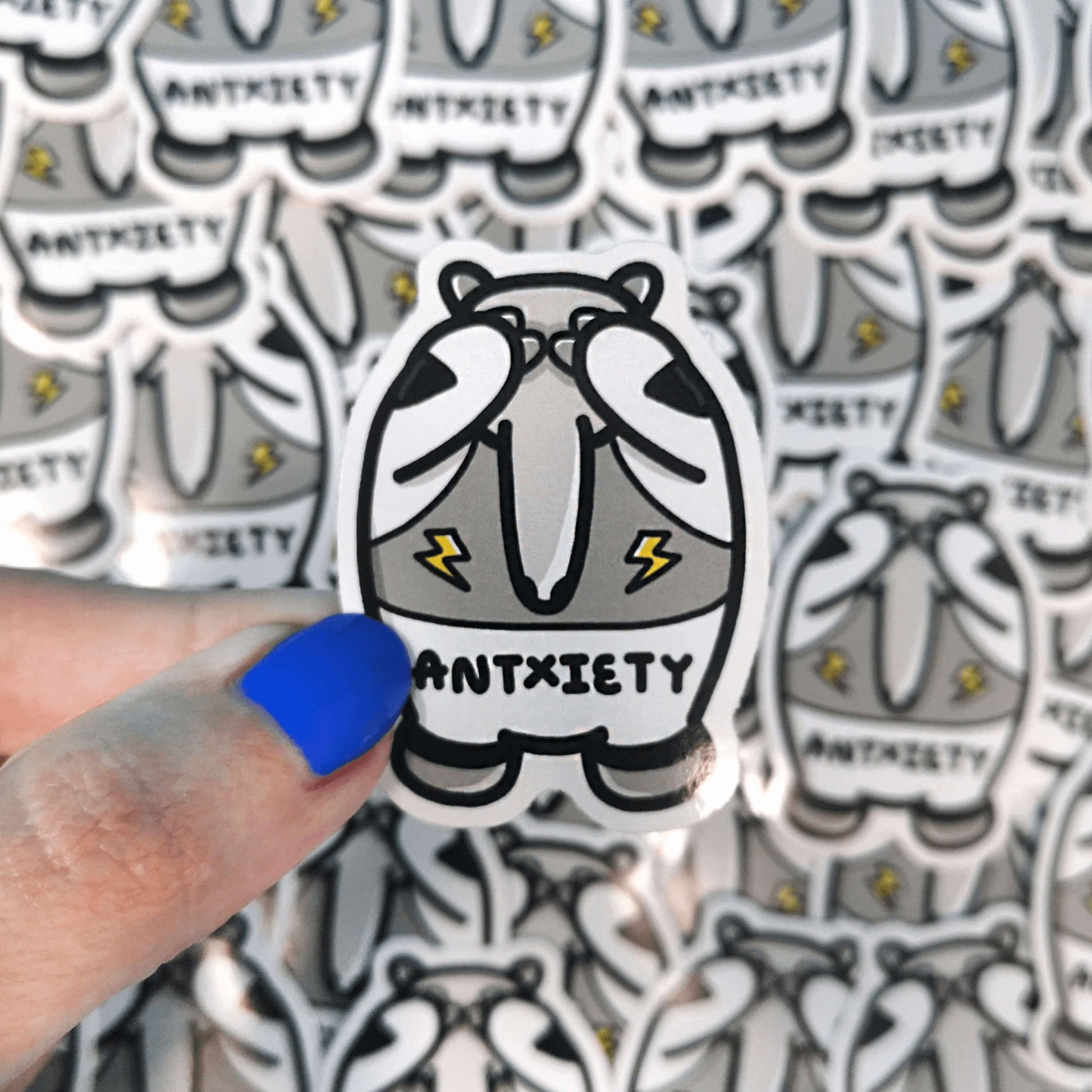 Antxiety Sticker Anxiety being held over lots of the stickers. The sticker is a grey and white anteater with yellow lightning bolts and the word antxiety across its belly. The sticker is designed to raise awareness for anxiety disorders and anxious emotions.