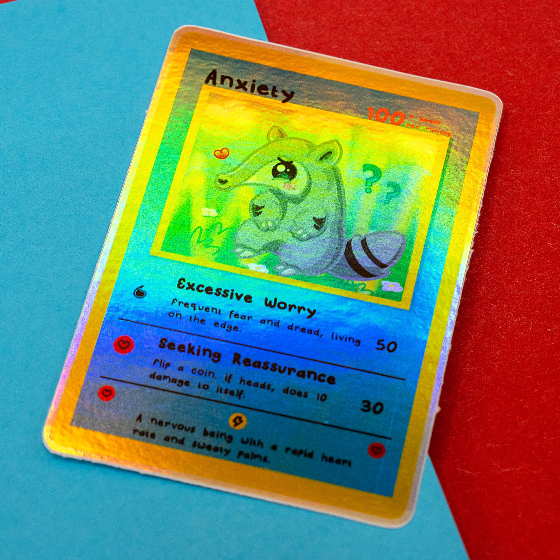 A holographic Innabox Neurodivergent Collectable Trading Card Sticker – Anxiety, showing the shiny rainbow effect across the anteater character artwork. The trading-card style sticker highlights traits like Excessive Worry and Seeking Reassurance. Photographed on a red and blue backdrop, perfect for collectors of disability-inspired stationery.