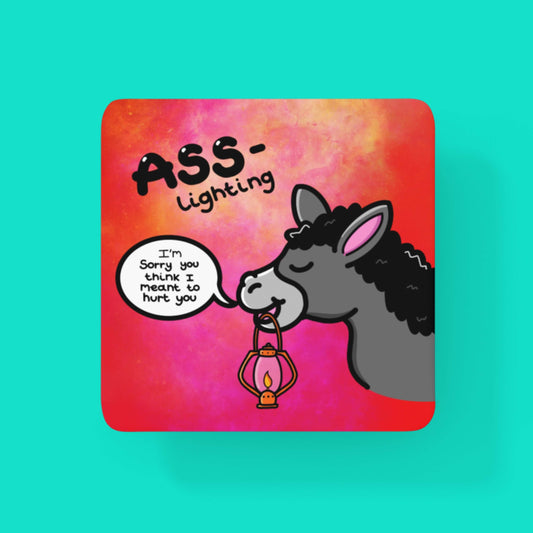 The Ass-Lighting Coaster - Gaslighting on a blue background. The red and pink gradient wooden coaster features a grey smiling donkey holding a lit orange lantern in its mouth with a speech bubble reading 'I'm sorry you think I meant to hurt you'. Above the donkey is black text that reads 'Ass-lighting'.