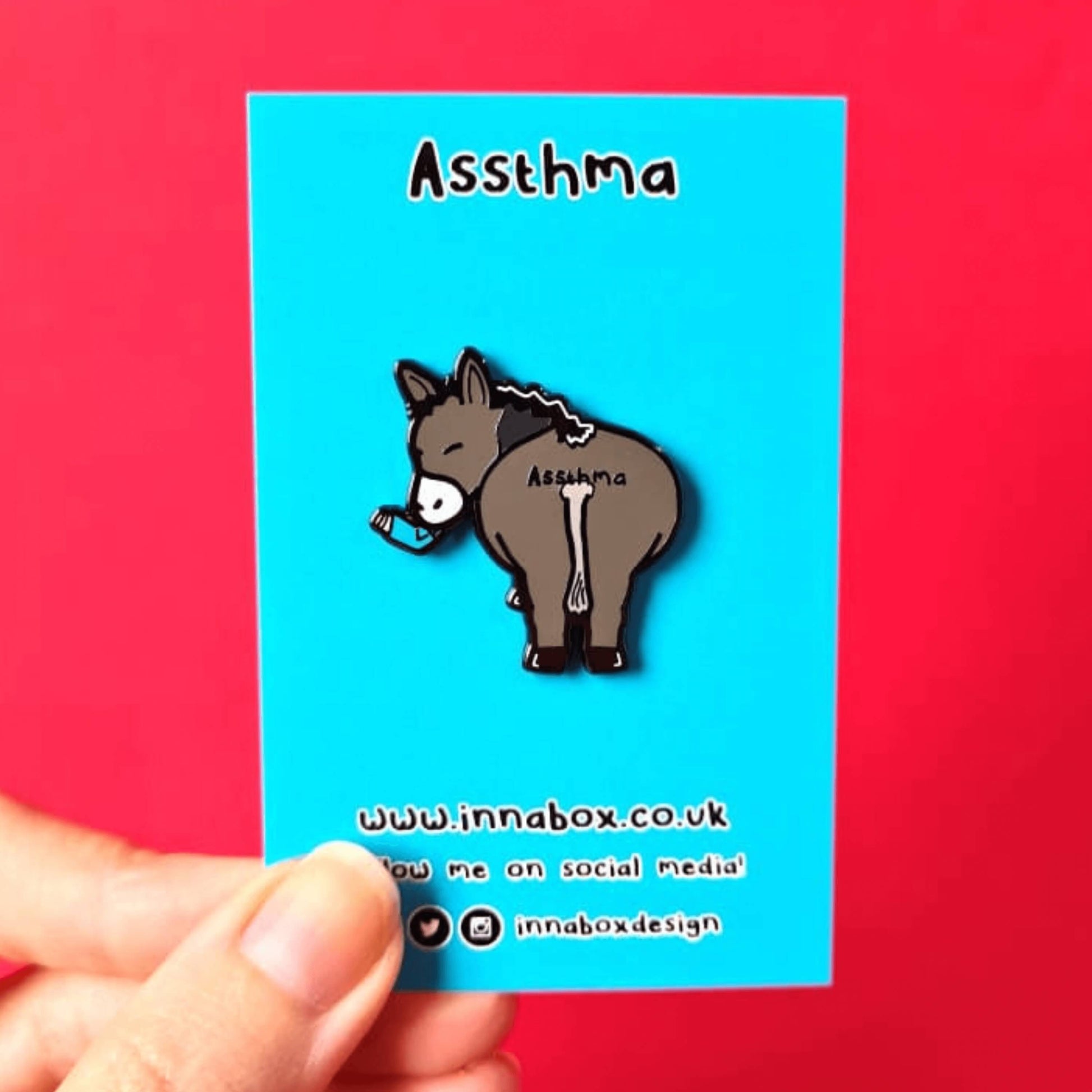 Assthma Enamel Pin - Asthma on blue backing card being held over a red background. A grey donkey ass showing its behind with a blue asthma pump in its mouth and the word Assthma across its behind. The pin is designed to raise awareness for asthma.