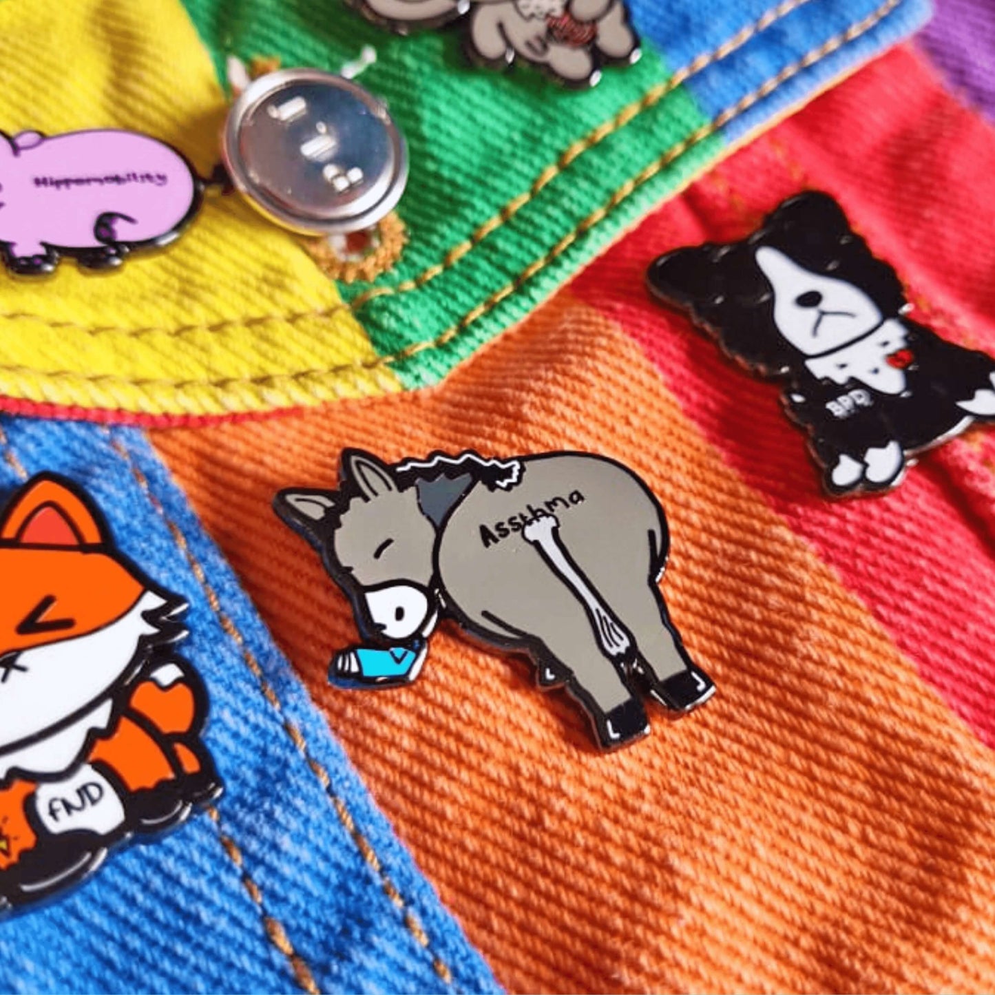 Assthma Enamel Pin - Asthma shown on a rainbow denim jacket with other innabox pins. A grey donkey ass showing its behind with a blue asthma pump in its mouth and the word Assthma across its behind. The pin is designed to raise awareness for asthma.
