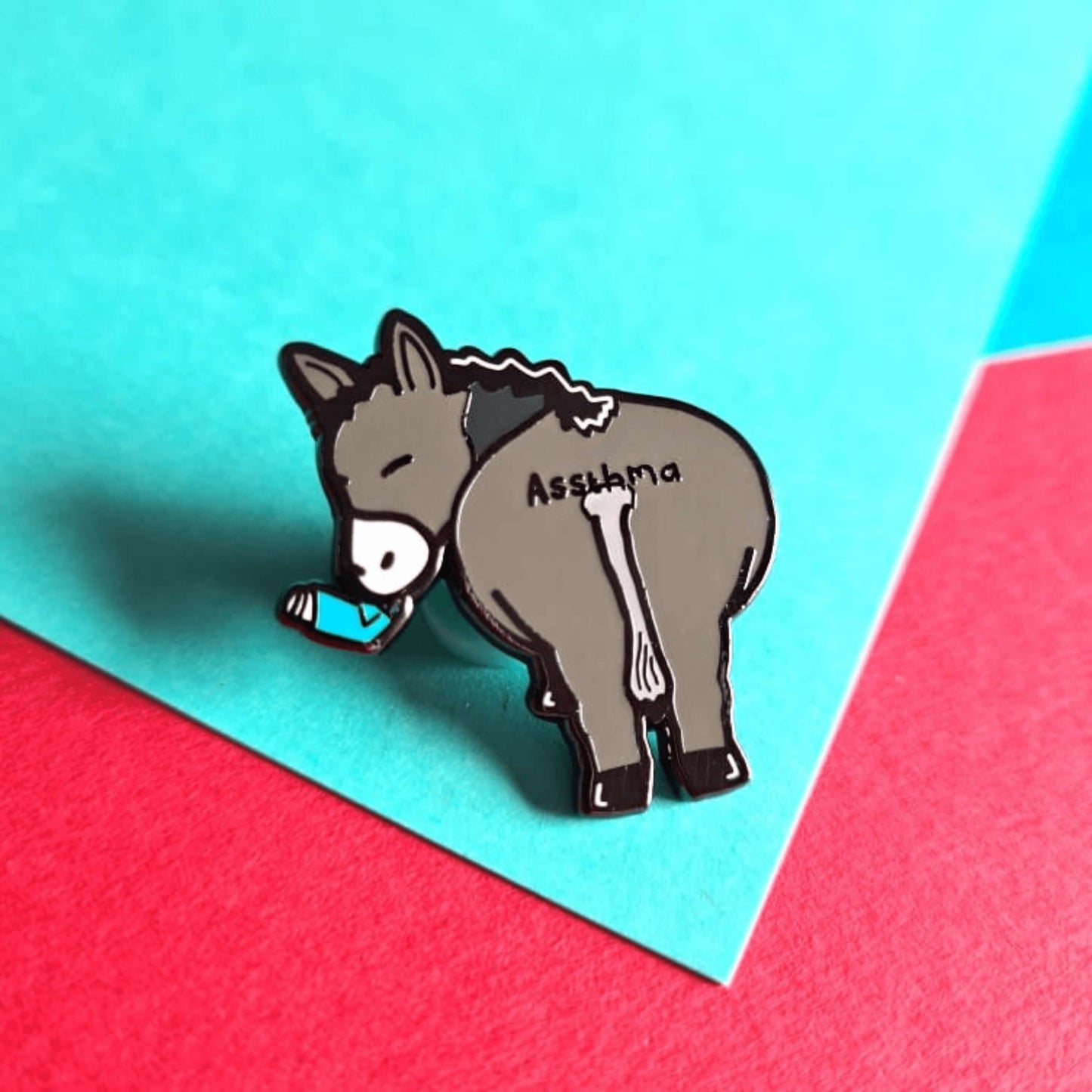Assthma Enamel Pin - Asthma on a blue, green and red background. A grey donkey ass showing its behind with a blue asthma pump in its mouth and the word Assthma across its behind. The pin is designed to raise awareness for asthma.