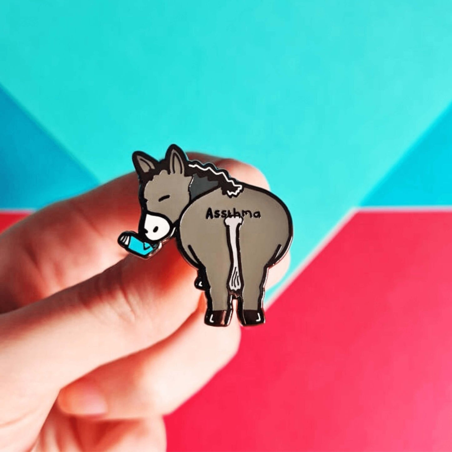Assthma Enamel Pin - Asthma being held over a blue, green and red background. A grey donkey ass showing its behind with a blue asthma pump in its mouth and the word Assthma across its behind. The pin is designed to raise awareness for asthma.