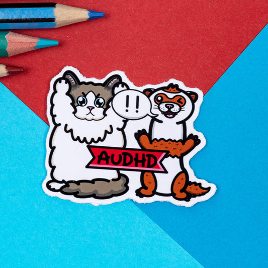 A sticker of a cat and a ferret with their arms in the air and a speech bubble with !! in it. It has AuDHD written on a red banner on the front. The sticker is on a red and blue background. The hand drawn design is raising awareness for neurodivergent people with adhd and autism combined.