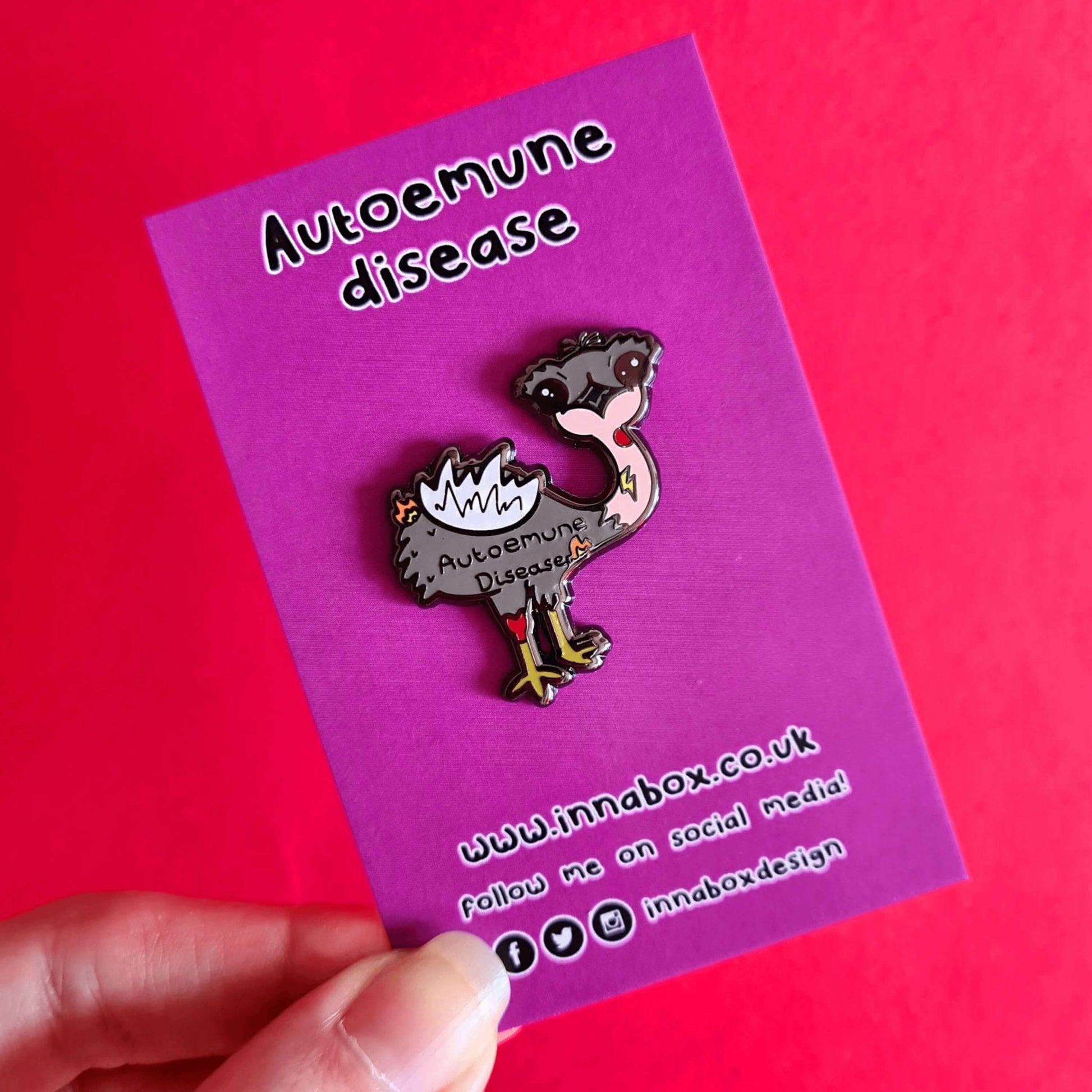 Autoemune Enamel Pin - Autoimmune Disease on a purple backing card being held over a red background. The pin is a grey and white emu bird with various problems highlighted with lightning bolts, flames and red circles with the words autoemune disease written across its belly. Design created to raise awareness for autoimmune diseases.