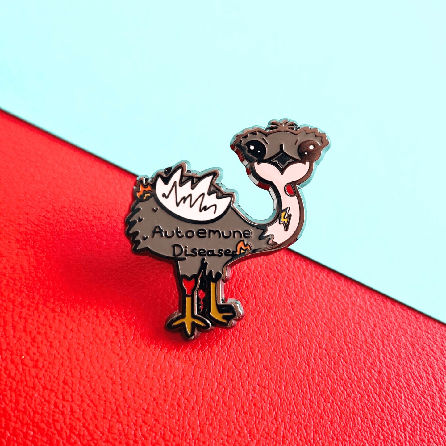 Autoemune Enamel Pin - Autoimmune Disease shown on a red and blue background. The pin is a grey and white emu bird with various problems highlighted with lightning bolts, flames and red circles with the words autoemune disease written across its belly. Design created to raise awareness for autoimmune diseases.