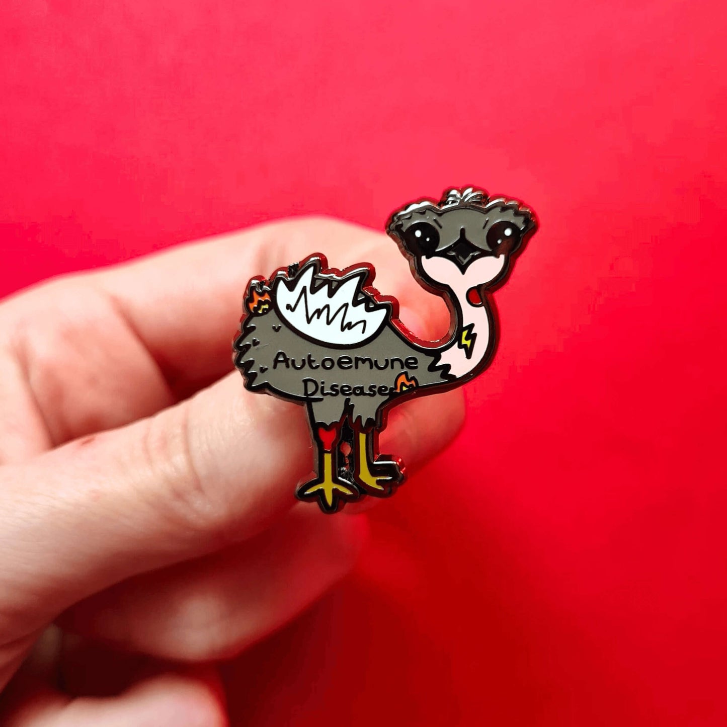 Autoemune Enamel Pin - Autoimmune Disease being held over a red background. The pin is a grey and white emu bird with various problems highlighted with lightning bolts, flames and red circles with the words autoemune disease written across its belly. Design created to raise awareness for autoimmune diseases.