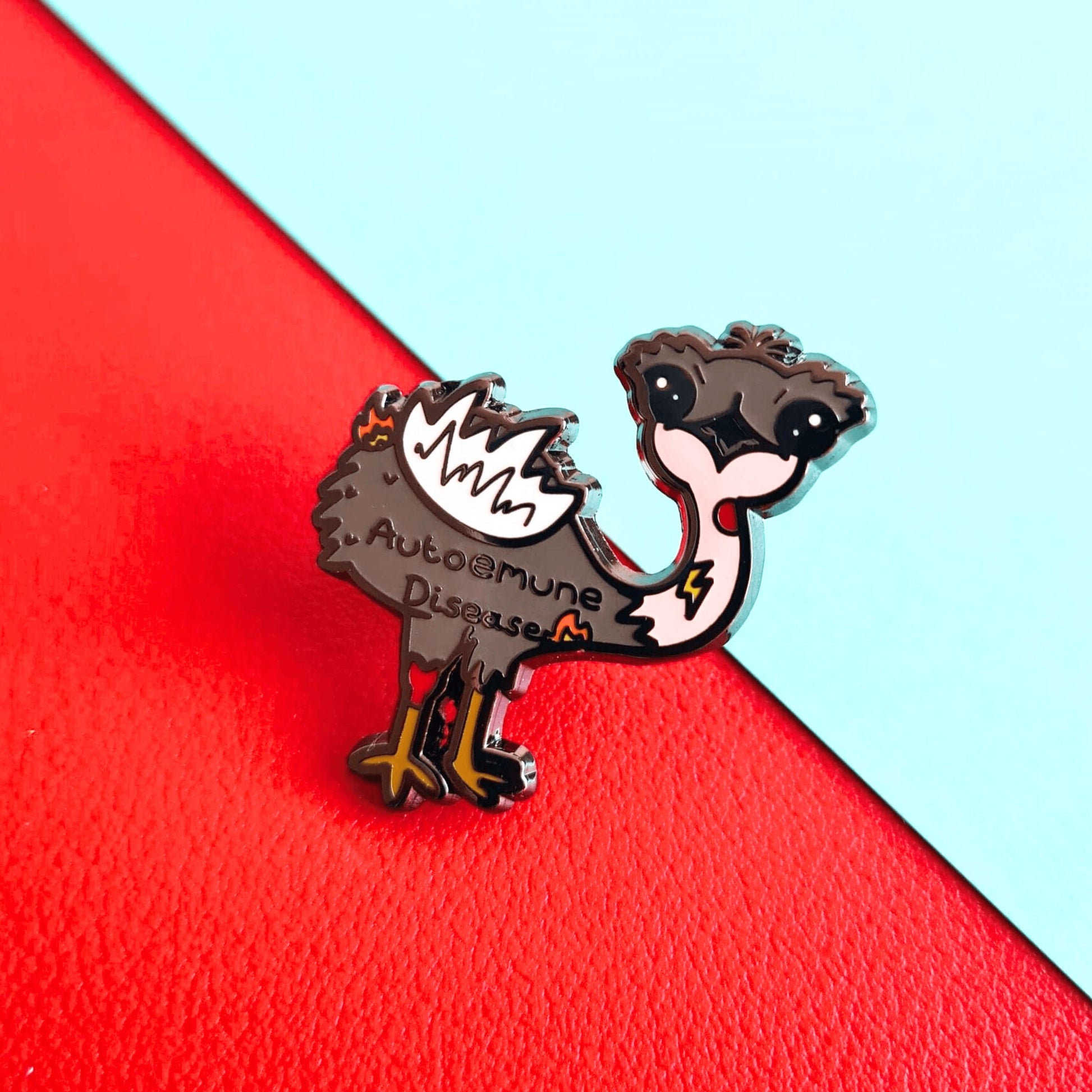 Autoemune Enamel Pin - Autoimmune Disease shown on a red and blue background. The pin is a grey and white emu bird with various problems highlighted with lightning bolts, flames and red circles with the words autoemune disease written across its belly. Design created to raise awareness for autoimmune diseases.