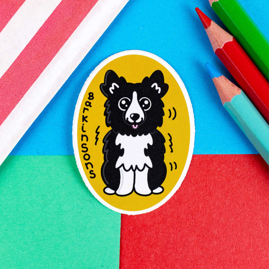 Barkinsons Disease Sticker - Parkinsons on a red, blue and green background with colouring pencils and a red stripe candy bag. The sticker is yellow and oval shaped with a black and white fluffy dog in the middle sat down with big sparkly eyes and pink tongue hanging out. There are black squiggles around the dog and 'Barkinsons' written in black vertical letters. The design is to raise awareness for parkinson's disease.