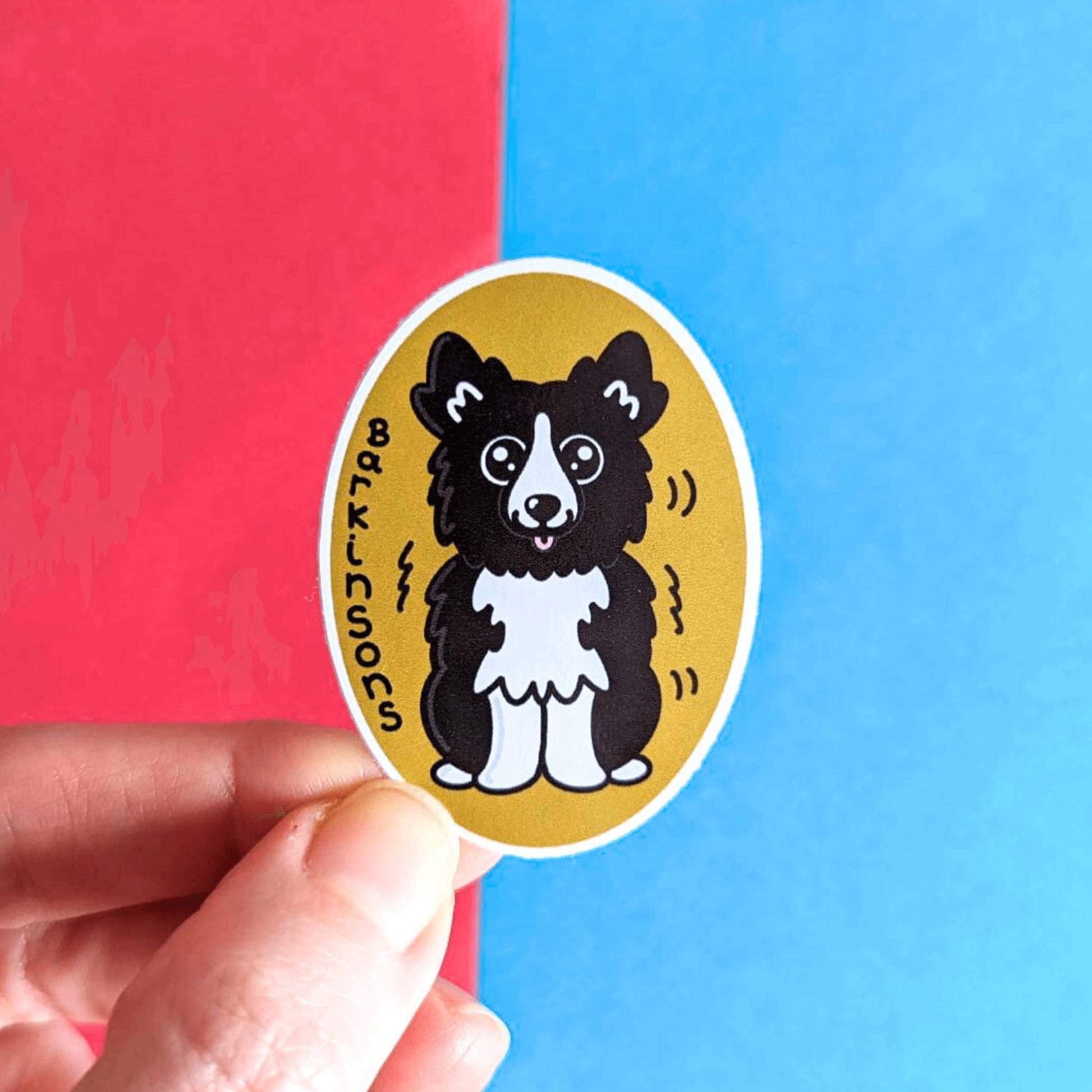 Barkinsons Disease Sticker - Parkinsons being held over a red and blue background. The sticker is yellow and oval shaped with a black and white fluffy dog in the middle sat down with big sparkly eyes and pink tongue hanging out. There are black squiggles around the dog and 'Barkinsons' written in black vertical letters. The design is to raise awareness for parkinson's disease.