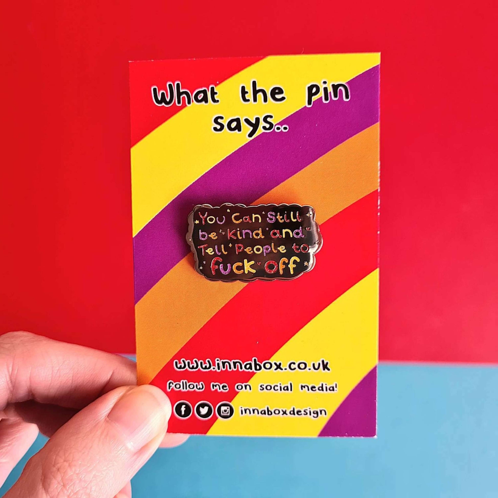 The You Can Still be Kind and Tell People to F**k Off Enamel Pin on rainbow backing card being held over a red and blue background. The silver plated enamel pin has rainbow writing reading 'you can still be kind and tell people to fuck off' with multicoloured sparkles, dots and stars.