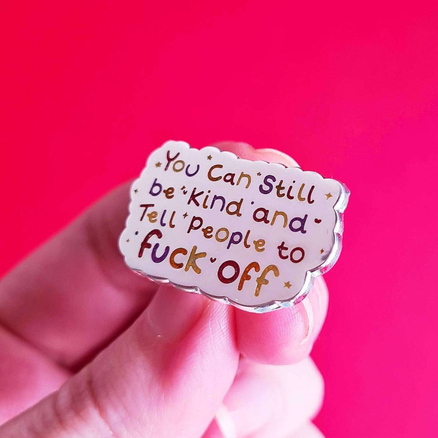 The You Can Still be Kind and Tell People to F**k Off Enamel Pin being held over a red background. The silver plated enamel pin has rainbow writing reading 'you can still be kind and tell people to fuck off' with multicoloured sparkles, dots and stars.