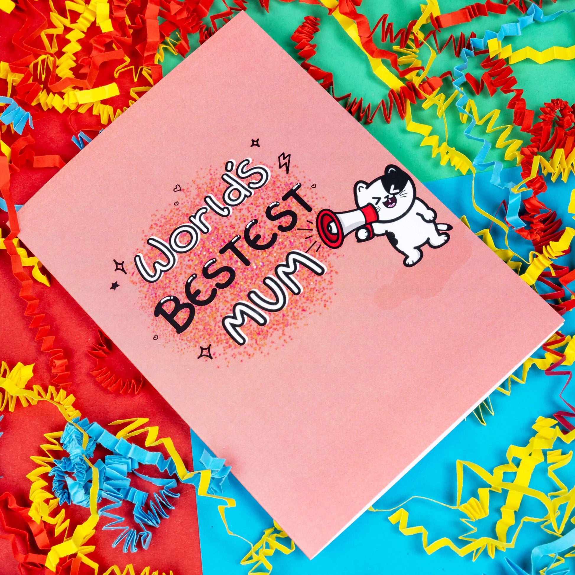 The World's Bestest Mum Cat Card on a red, blue and green background with red, yellow and blue crinkle card confetti. The pink a6 mother's day card features pink, orange and yellow dotwork background with white and black doodle text reading 'world's bestest mum'. Next to the text is a smiling black and white cat shouting through a megaphone.