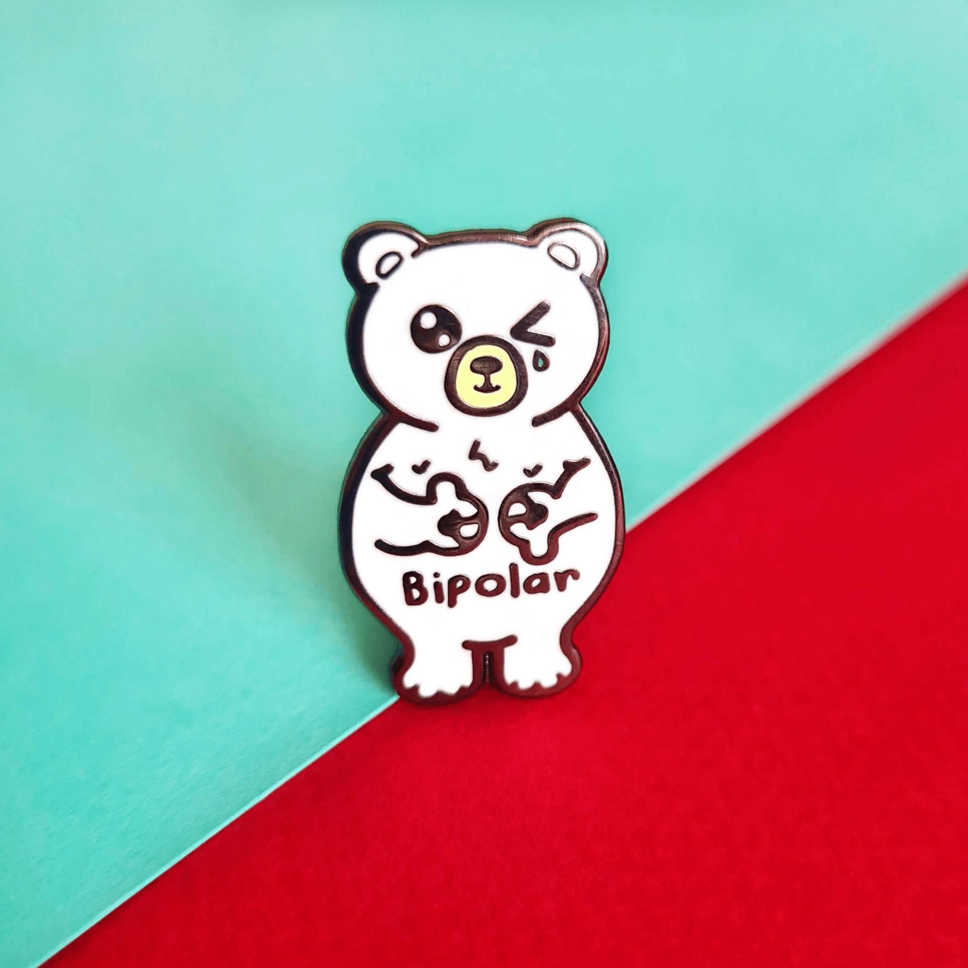 Bipolar Bear Enamel Pin - Bipolar on a red and blue background. The pin is a white polar bear with one eye closed crying whilst smiling clutching its chest, across its tummy reads bipolar. The pun pin is raising awareness for bipolar disorder.