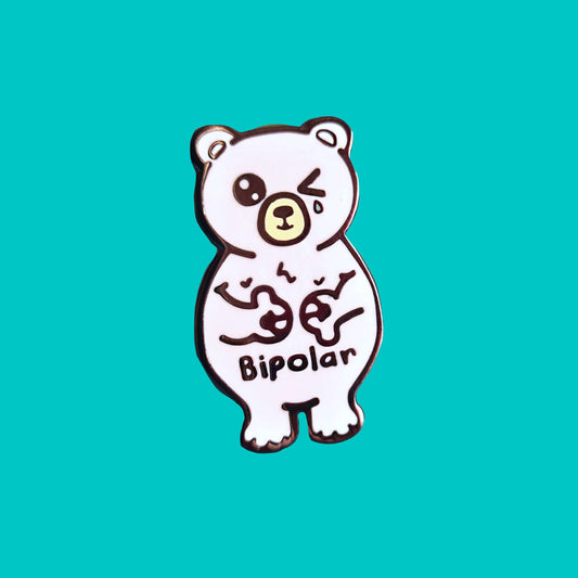 Bipolar Bear Enamel Pin - Bipolar on a blue background. The pin is a white polar bear with one eye closed crying whilst smiling clutching its chest, across its tummy reads bipolar. The pun pin is raising awareness for bipolar disorder.