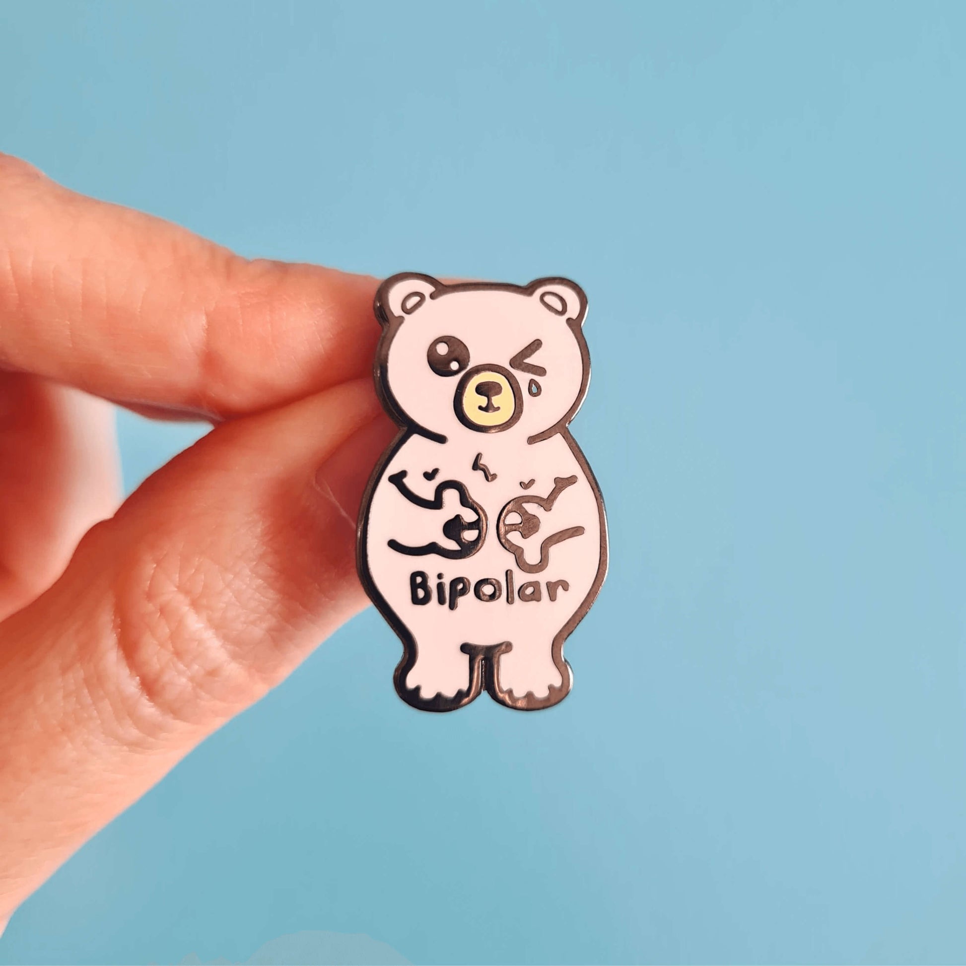 Bipolar Bear Enamel Pin - Bipolar being held over a blue background. The pin is a white polar bear with one eye closed crying whilst smiling clutching its chest, across its tummy reads bipolar. The pun pin is raising awareness for bipolar disorder.