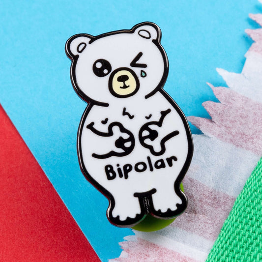 Bipolar Bear Enamel Pin - Bipolar on a blue and red card background. The pin is a white polar bear with one eye closed crying whilst smiling clutching its chest, across its tummy reads bipolar. The pun pin is raising awareness for bipolar disorder.