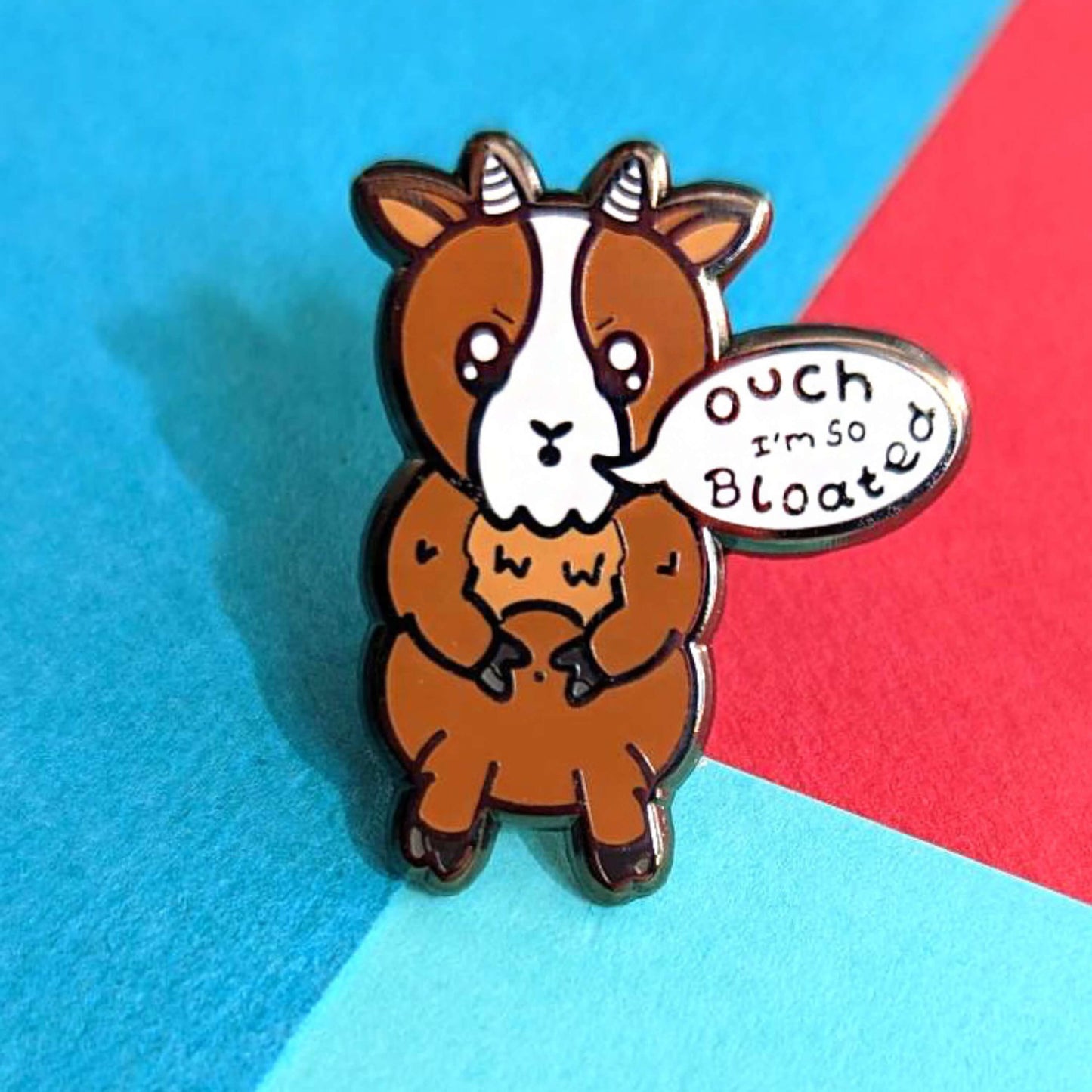 The Bloat Goat Enamel Pin on a red and blue background. The brown frustrated goat pin badge is stood up clutching its tummy with a speech bubble reading 'ouch I'm so bloated'. The hand drawn design is raising awareness for chronic illnesses or hidden disabilities that cause bloating.