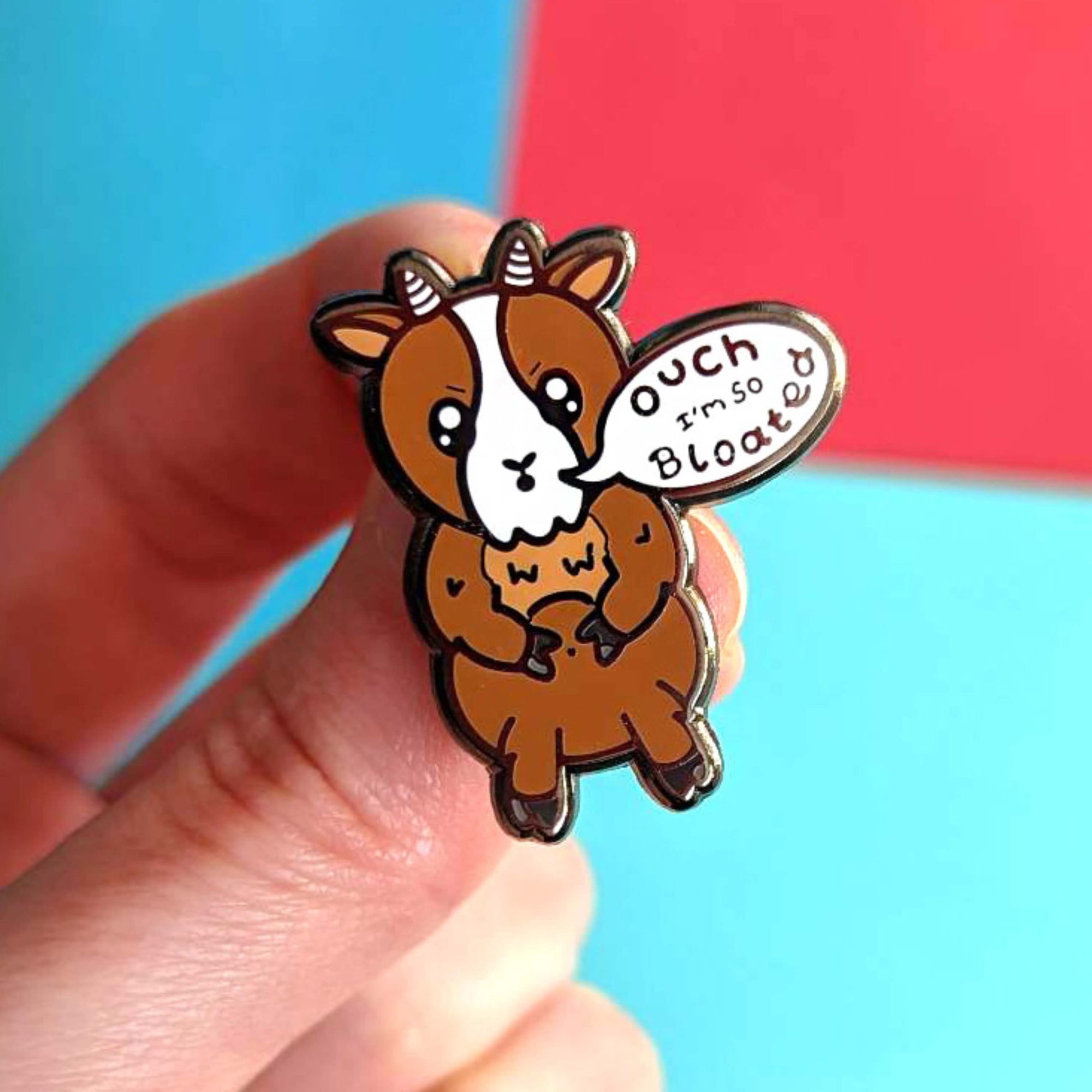 The Bloat Goat Enamel Pin held over a red and blue background. The brown frustrated goat pin badge is stood up clutching its tummy with a speech bubble reading 'ouch I'm so bloated'. The hand drawn design is raising awareness for chronic illnesses or hidden disabilities that cause bloating.