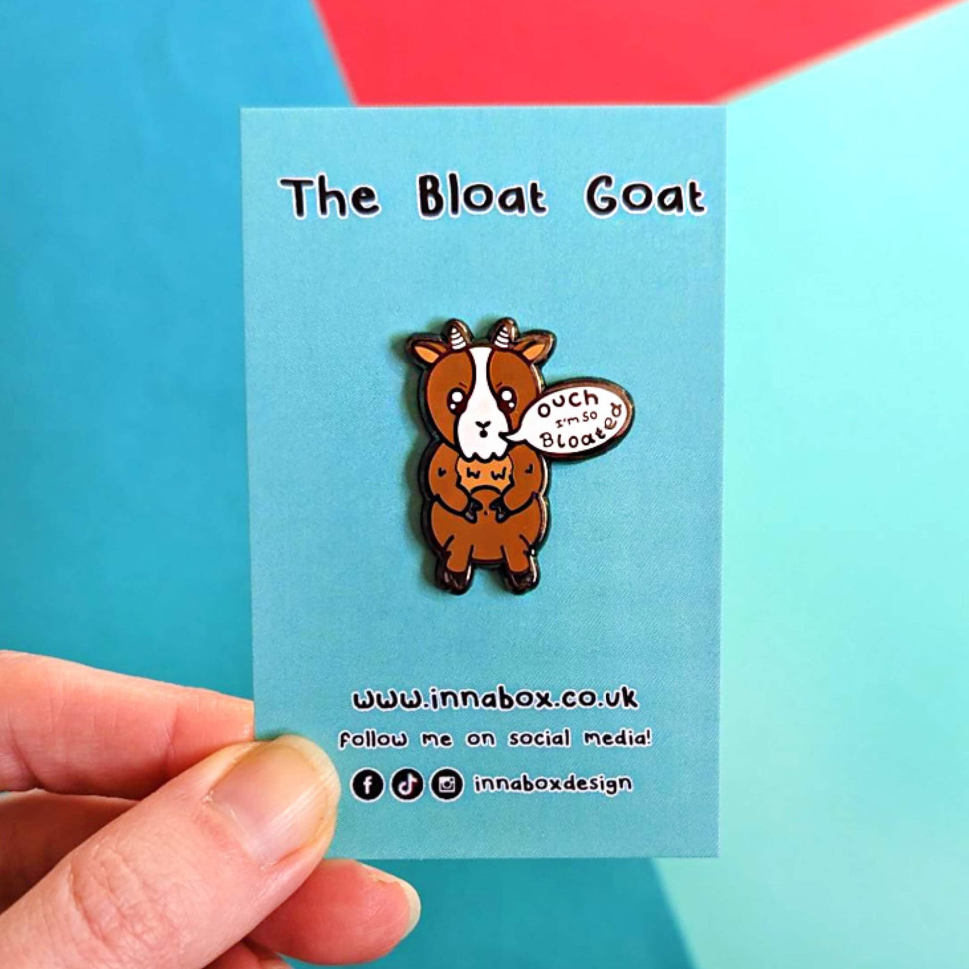 The Bloat Goat Enamel Pin on its blue backing card being held over a blue and red background. The brown frustrated goat pin badge is stood up clutching its tummy with a speech bubble reading 'ouch I'm so bloated'. The hand drawn design is raising awareness for chronic illnesses or hidden disabilities that cause bloating.
