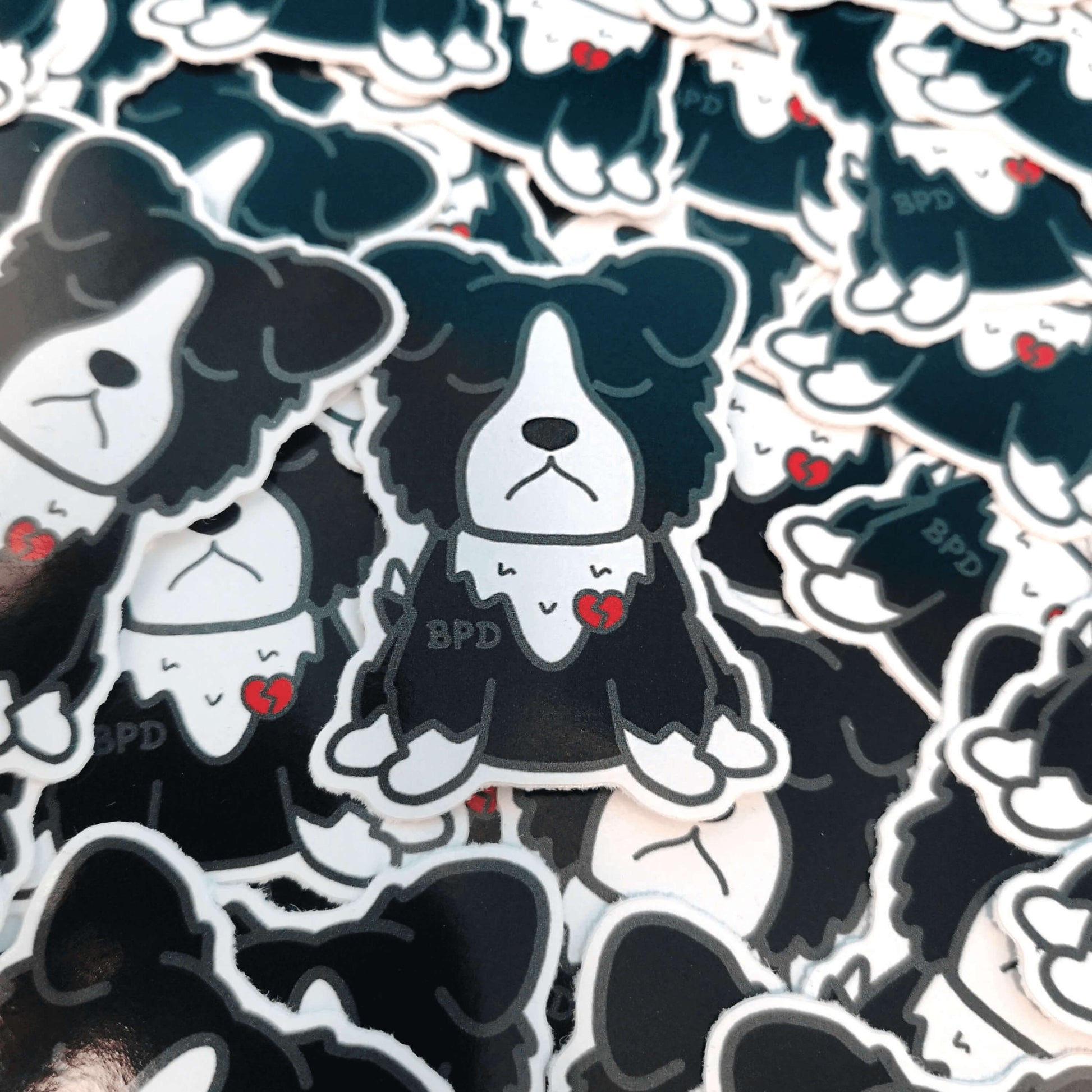 BPD Sticker - Borderline Personality Disorder laid amongst a pile of other bpd dog stickers. The black and white sad fluffy dog sticker has a broken heart with BPD written across its chest. The sticker is designed to raise awareness for Borderline Personality Disorder.