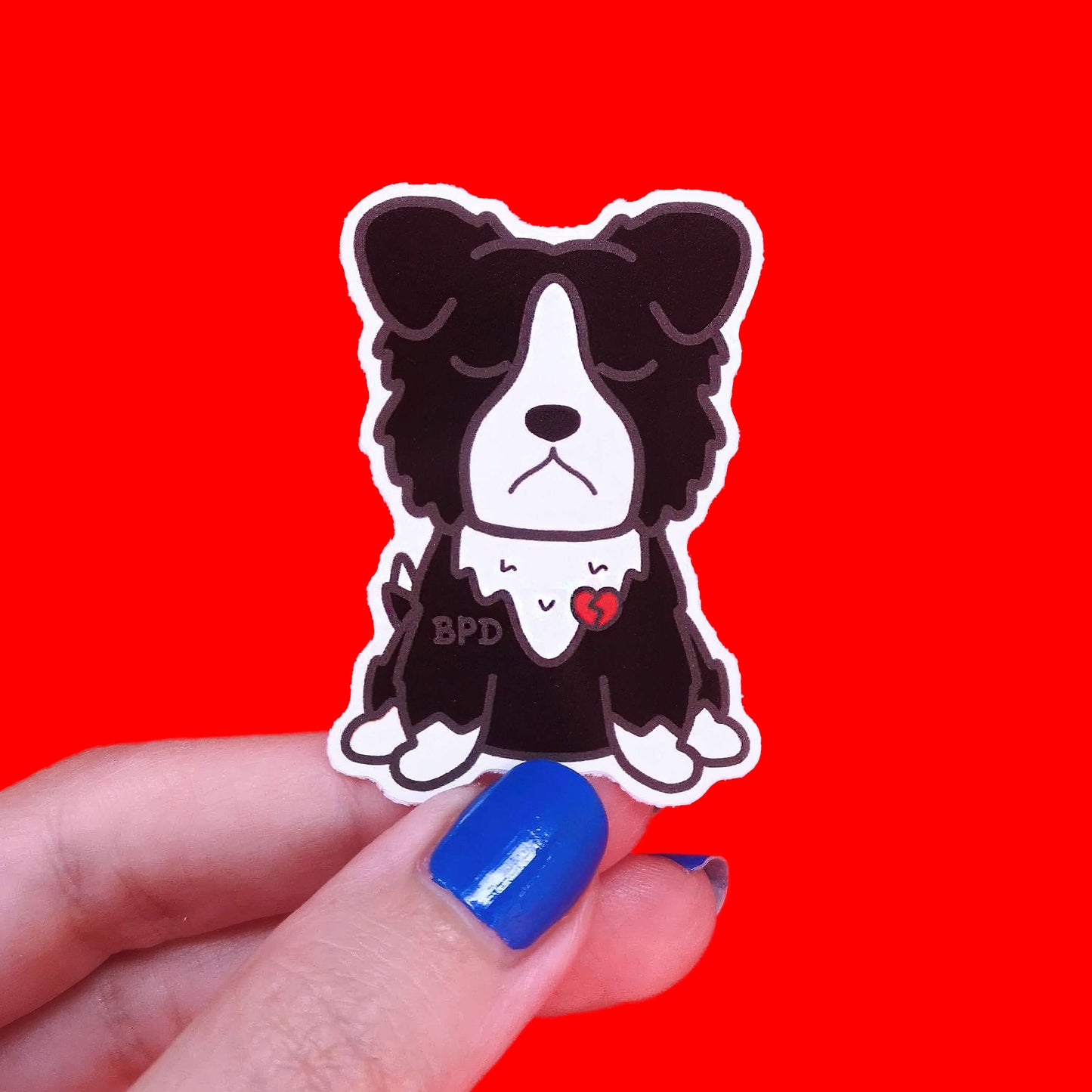 BPD Sticker - Borderline Personality Disorder being held up by a hand with blue nail varnish over a red background. The black and white sad fluffy dog sticker has a broken heart with BPD written across its chest. The sticker is designed to raise awareness for Borderline Personality Disorder.