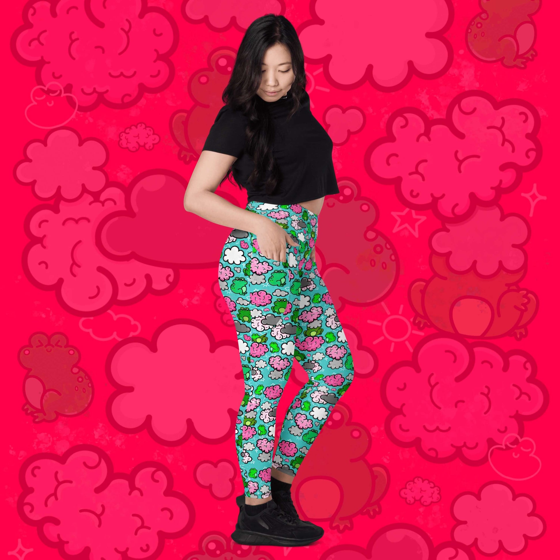 The Brain Frog Leggings with pockets - Brain Fog being worn by a model facing right with one leg bent, one hand in the pocket and looking down on a red background with a faded brain frog print. The leggings are an aqua blue base with various green kawaii face frogs with pink blush cheeks, pink brain style clouds, white and grey clouds and white sparkles all over. The design was created to raise awareness of Brain Fog.