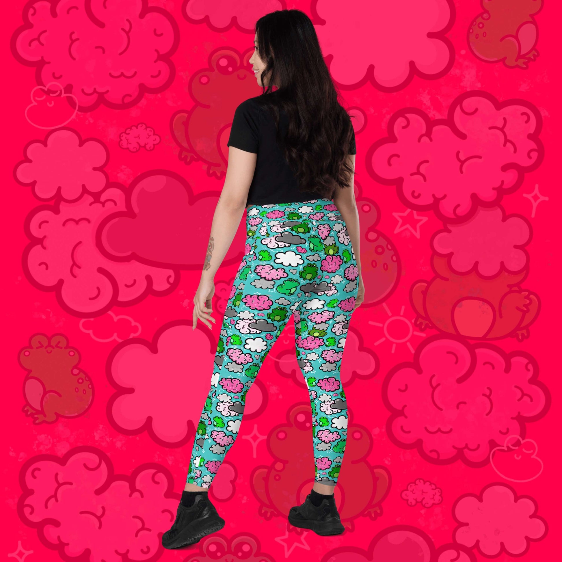 The Brain Frog Leggings with pockets - Brain Fog being worn by a model facing away with one leg stretched out to the side and smiling looking slightly over their shoulder on a red background with a faded brain frog print. The leggings are an aqua blue base with various green kawaii face frogs with pink blush cheeks, pink brain style clouds, white and grey clouds and white sparkles all over. The design was created to raise awareness of Brain Fog.