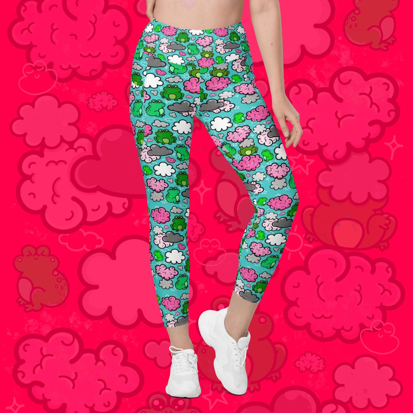 The Brain Frog Leggings with pockets - Brain Fog being worn by a model facing forward with their right leg bent on a red background with a faded brain frog print, the photo is cropped from the hips down. The leggings are an aqua blue base with various green kawaii face frogs with pink blush cheeks, pink brain style clouds, white and grey clouds and white sparkles all over. The design was created to raise awareness of Brain Fog.