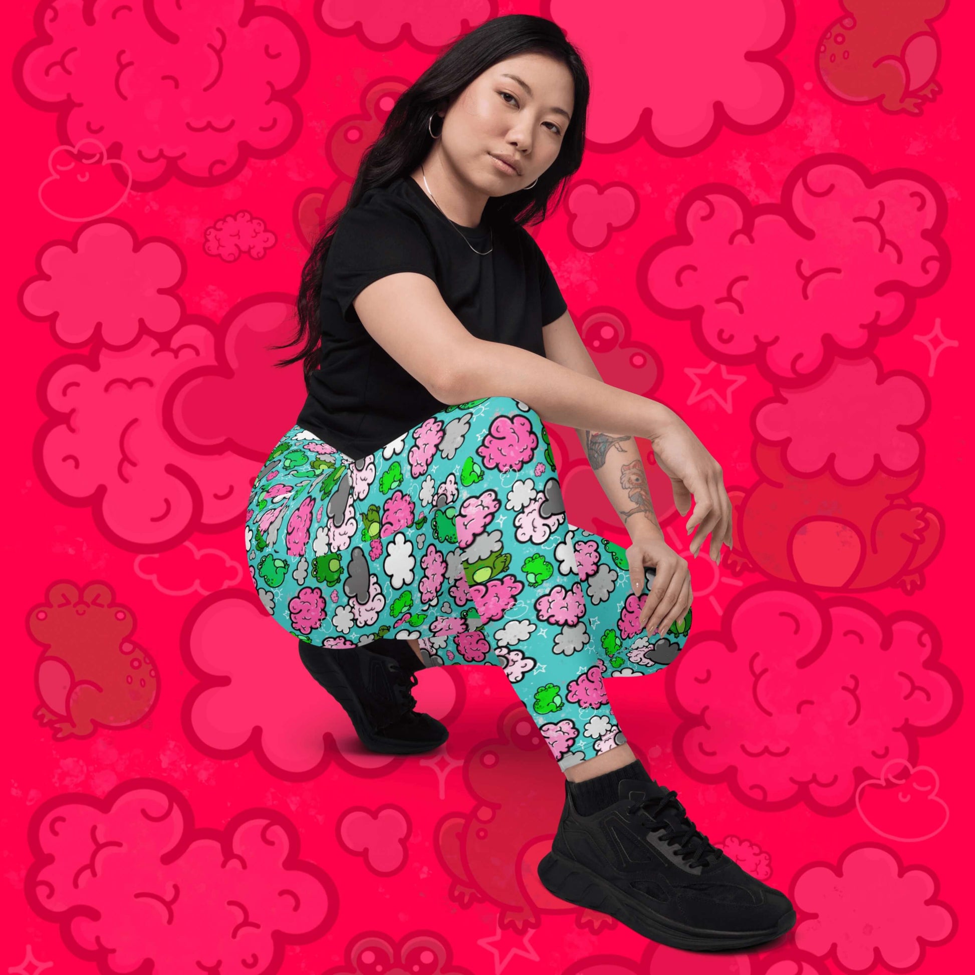 The Brain Frog Leggings with pockets - Brain Fog being worn by a model facing forward crouched down with their arms resting on their legs whilst smising to camera on a red background with a faded brain frog print. The leggings are an aqua blue base with various green kawaii face frogs with pink blush cheeks, pink brain style clouds, white and grey clouds and white sparkles all over. The design was created to raise awareness of Brain Fog.