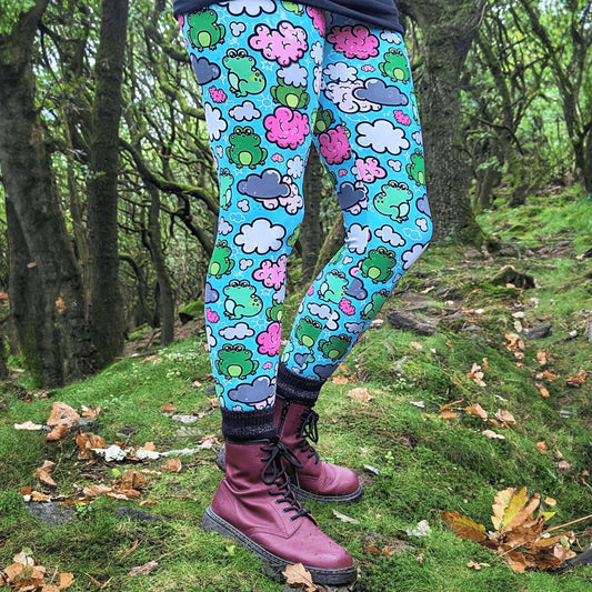 The Brain Frog Leggings - Brain Fog being worn by a model outside in a mossy tree forest area with red boots, sparkly black socks and a black top, the photo is cropped from the hips down. The leggings are an aqua blue base with various green kawaii face frogs with pink blush cheeks, pink brain style clouds, white and grey clouds and white sparkles all over. The design was created to raise awareness of Brain Fog.