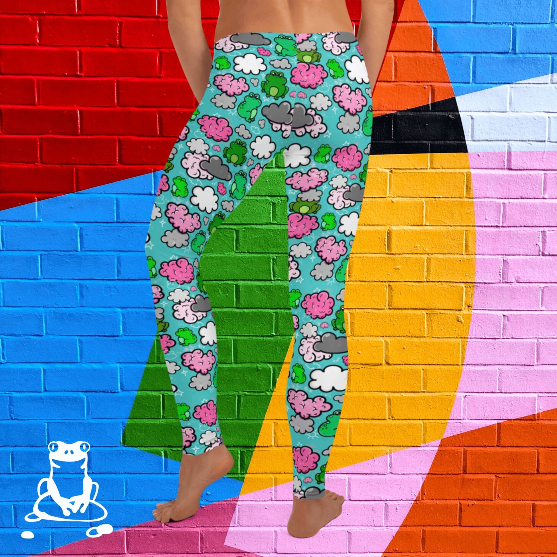 The Brain Frog Leggings - Brain Fog being worn by a model facing away with their left leg bent on a rainbow brick background, the photo is cropped from the hips down. The leggings are an aqua blue base with various green kawaii face frogs with pink blush cheeks, pink brain style clouds, white and grey clouds and white sparkles all over. The design was created to raise awareness of Brain Fog.