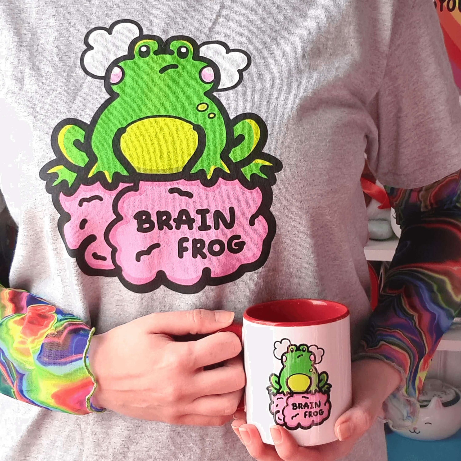 Close up of Nikky Box from innabox wearing a rainbow hair tie, a brain frog tshirt with a rainbow tie dye mesh long sleeve underneath with a Brain Frog Mug - Brain Fog resting on her front. The white mug has a red handle and inside with a front print of a confused frog with pink blush cheeks and two grey clouds above its head. Its sat on a pink brain with text that reads Brain Frog. The design was created to raise awareness for brain fog.