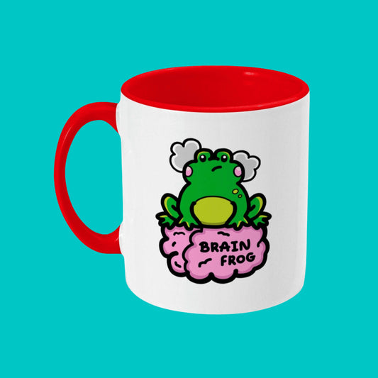 The Brain Frog Mug - Brain Fog on a blue background. The white mug has a red handle and inside with a front print of a confused frog with pink blush cheeks and two grey clouds above its head. Its sat on a pink brain with text that reads Brain Frog. The design was created to raise awareness for brain fog.
