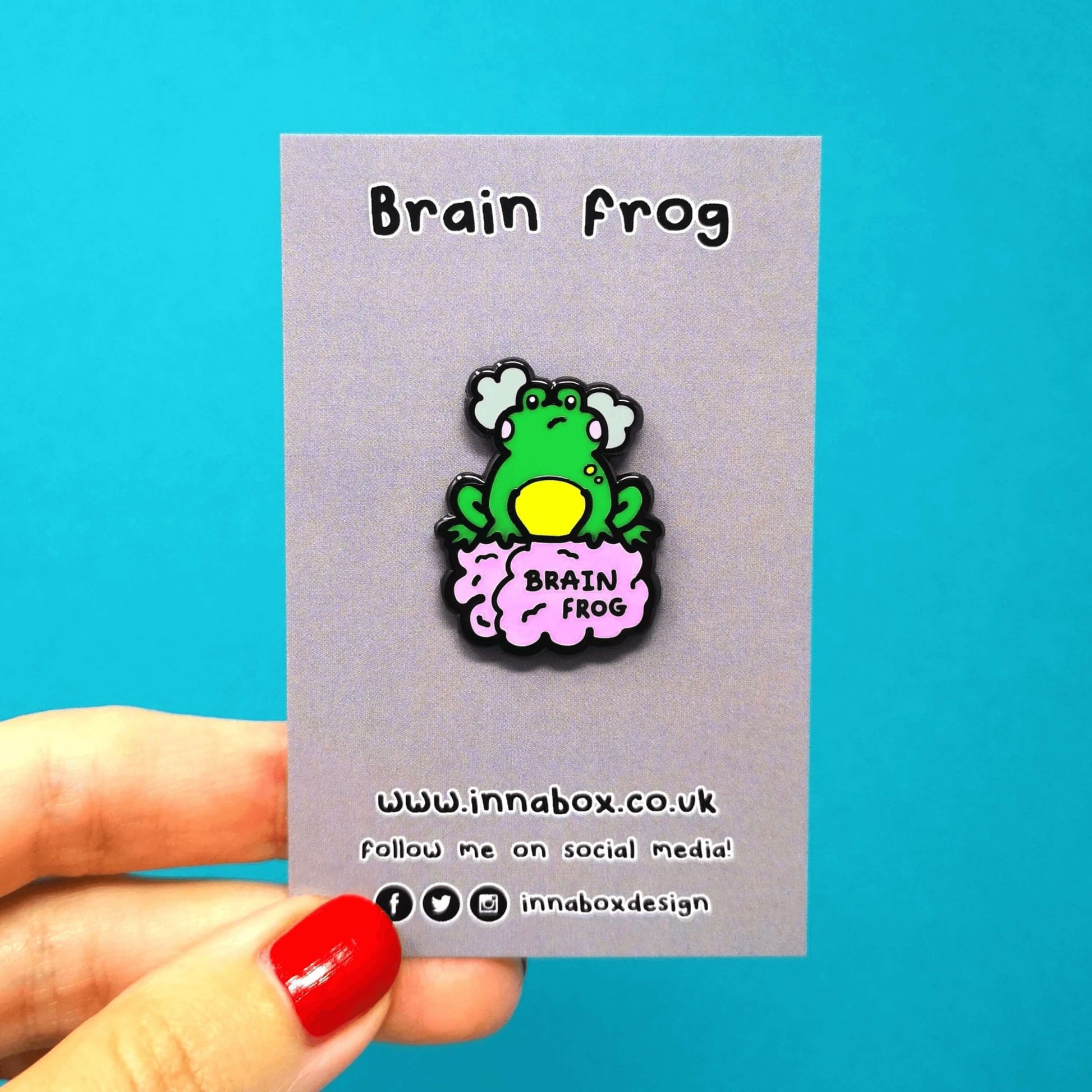 The Brain Frog Enamel Pin - Brain Fog on a grey backing card with the product name above and the innabox social media handles at the bottom, it is being held over a blue background. The pin is of a green frog with pink blush cheeks looking confused with two grey clouds by its head, it is sat on a pink brain with the text reading Brain Frog. The design is raising awareness for brain fog.