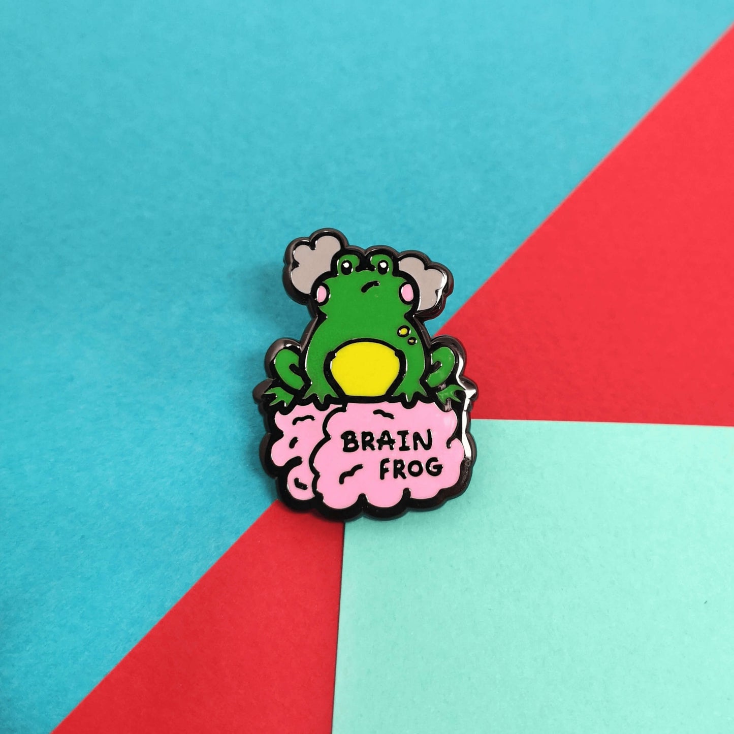 The Brain Frog Enamel Pin - Brain Fog on a red, green and blue background. The pin is of a green frog with pink blush cheeks looking confused with two grey clouds by its head, it is sat on a pink brain with the text reading Brain Frog. The design is raising awareness for brain fog.