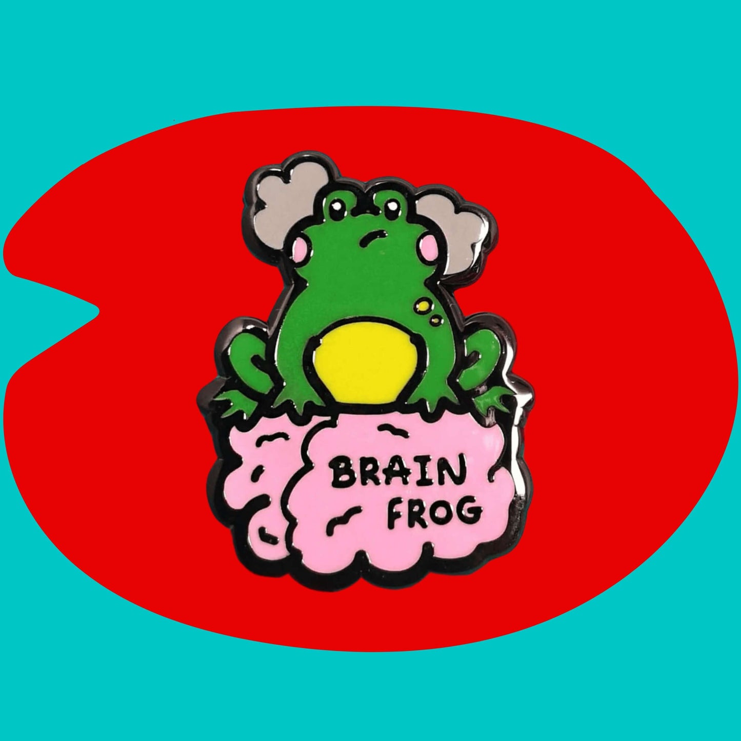 The Brain Frog Enamel Pin - Brain Fog on a blue and red background. The pin is of a green frog with pink blush cheeks looking confused with two grey clouds by its head, it is sat on a pink brain with the text reading Brain Frog. The design is raising awareness for brain fog.