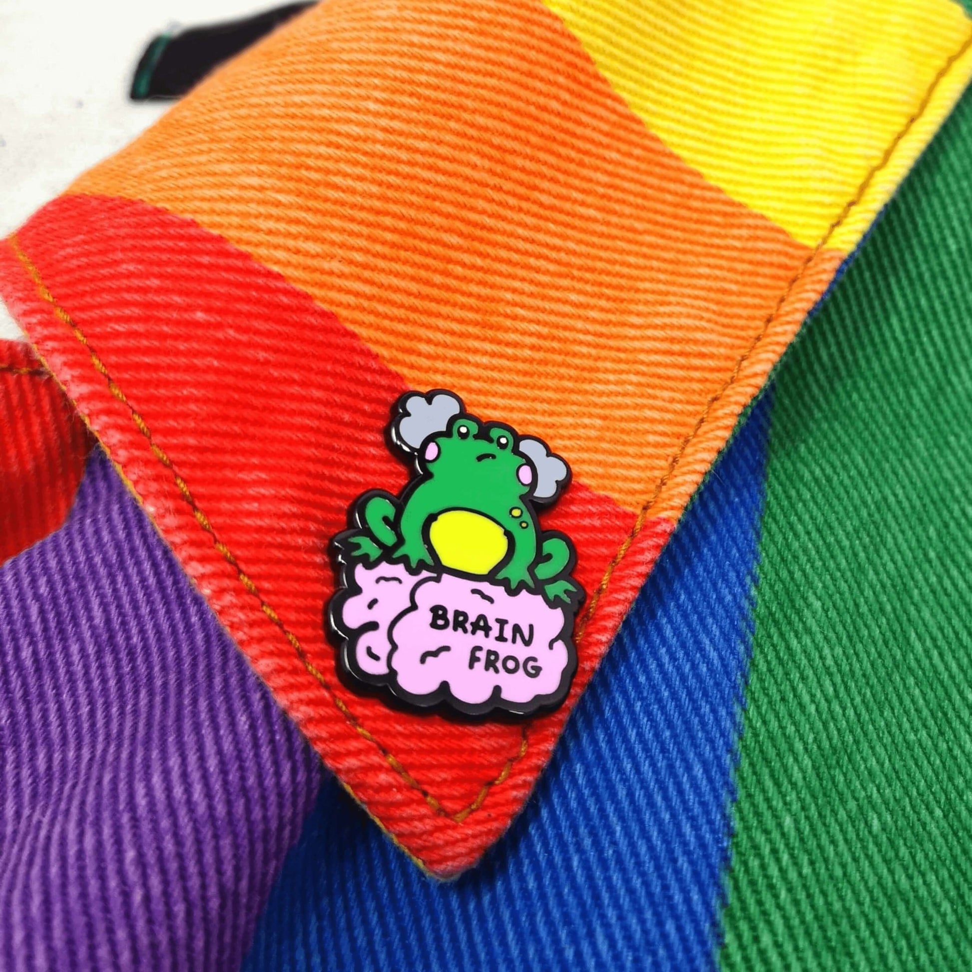 The Brain Frog Enamel Pin - Brain Fog on a rainbow denim jacket collar. The pin is of a green frog with pink blush cheeks looking confused with two grey clouds by its head, it is sat on a pink brain with the text reading Brain Frog. The design is raising awareness for brain fog.
