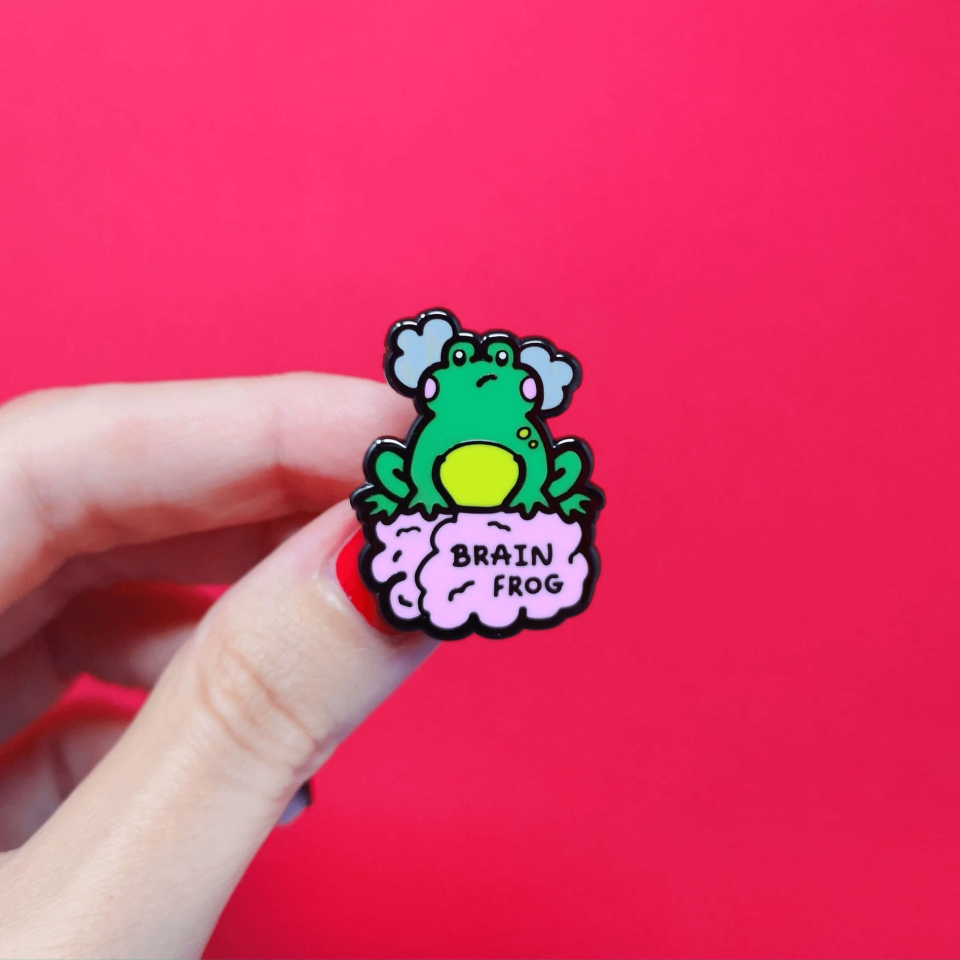 The Brain Frog Enamel Pin - Brain Fog being held over a red background. The pin is of a green frog with pink blush cheeks looking confused with two grey clouds by its head, it is sat on a pink brain with the text reading Brain Frog. The design is raising awareness for brain fog.