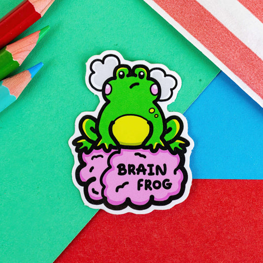 The Brain Frog Sticker - Brain Fog on a green, red and blue background with colouring pencils and a red stripe candy bag. The sticker is a confused frog with pink blush cheeks and two grey clouds above its head. Its sat on a pink brain with text that reads Brain Frog. The design was created to raise awareness for brain fog.