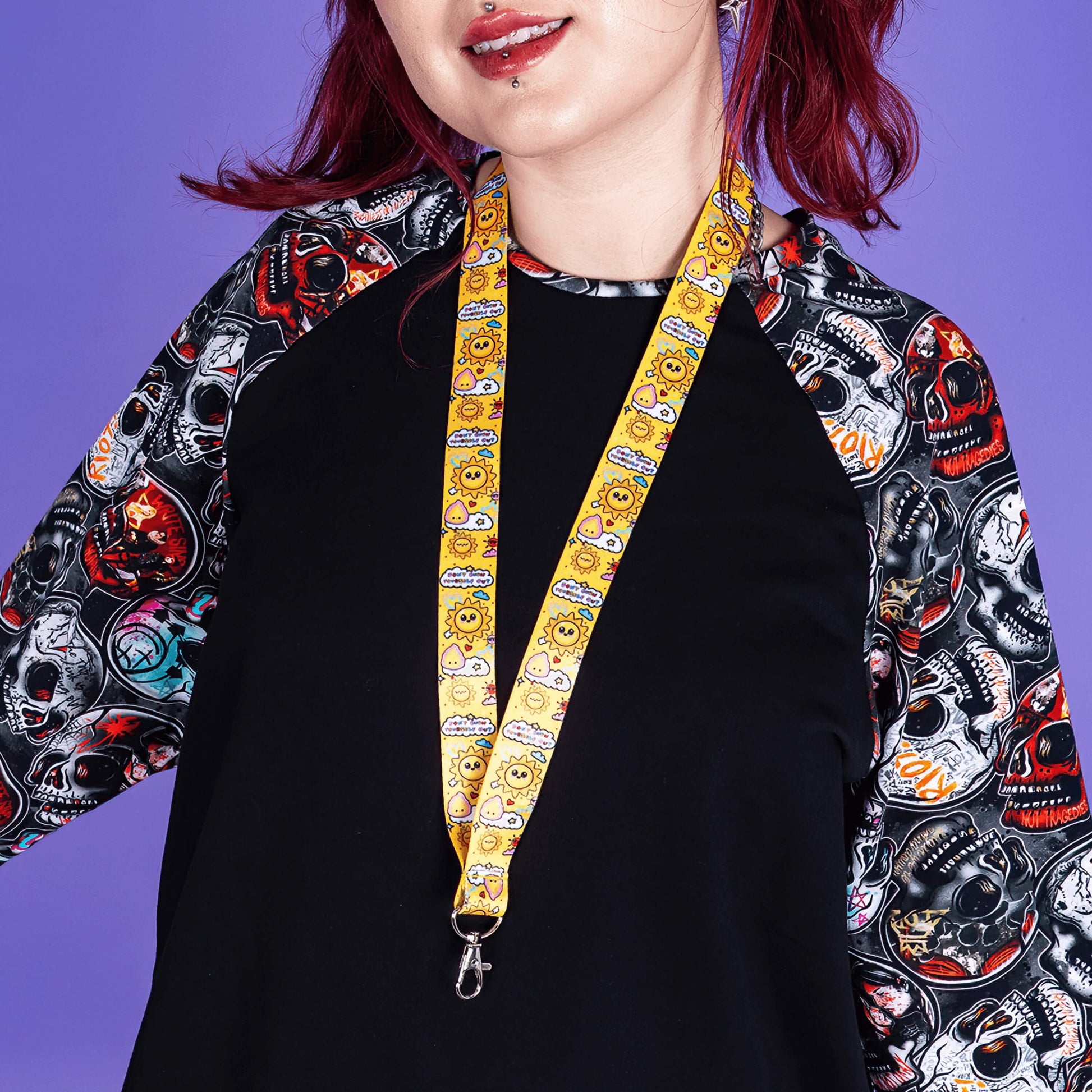 The Don't Burn Yourself Out Lanyard being worn by a red haired model wearing a wilde mode top in front of a purple background whilst smiling. Photo is cropped from the their lips to their waist. The lanyard is yellow with various kawaii face sunshines, sparkles, hearts, pink flames and pink, blue and white clouds with don't burn yourself out inside in rainbow. Design inspired by the self care movement.