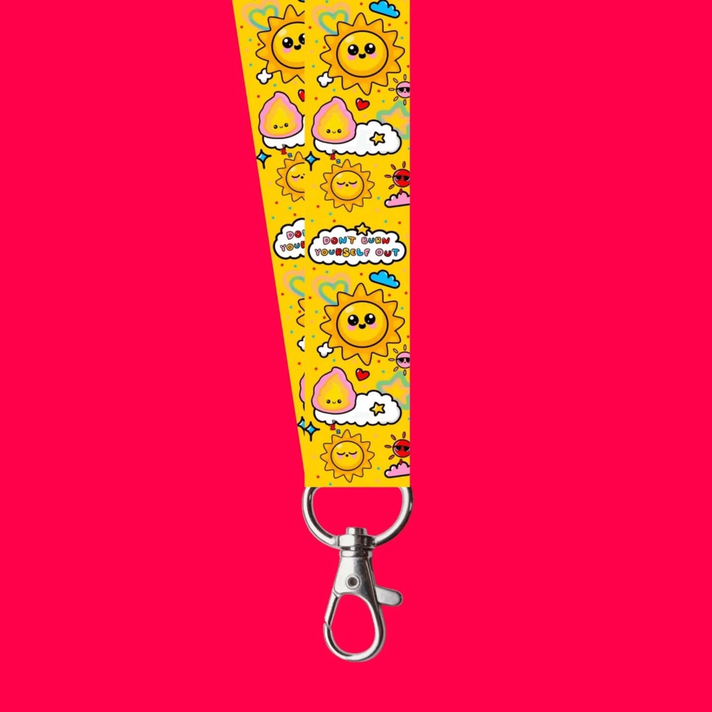 The Don't Burn Yourself Out Lanyard on a red background. The lanyard is yellow with various kawaii face sunshines, sparkles, hearts, pink flames and pink, blue and white clouds with don't burn yourself out inside in rainbow. Design inspired by the self care movement.