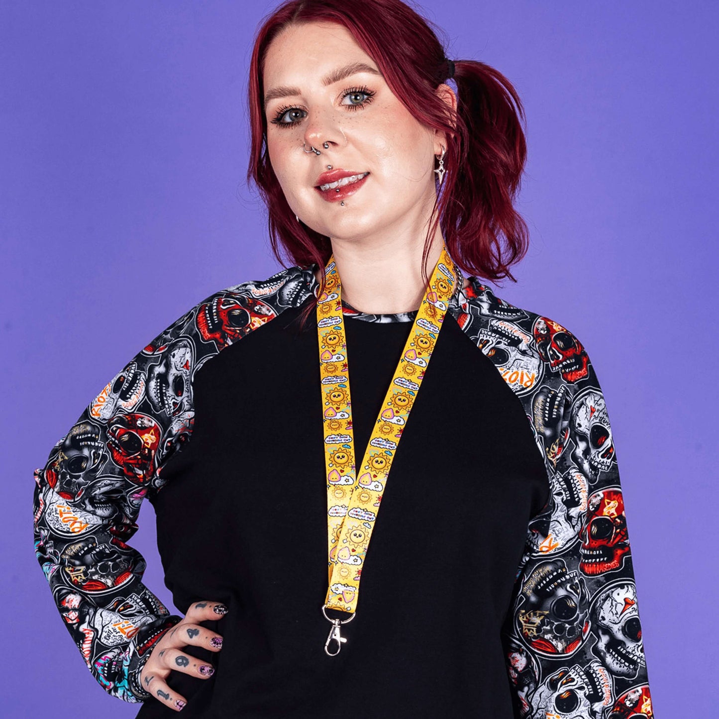 The Don't Burn Yourself Out Lanyard being worn by a red haired model wearing a wilde mode top in front of a purple background whilst smiling with their hand on their hip. Photo is cropped from the their head to their waist. The lanyard is yellow with various kawaii face sunshines, sparkles, hearts, pink flames and pink, blue and white clouds with don't burn yourself out inside in rainbow. Design inspired by the self care movement.