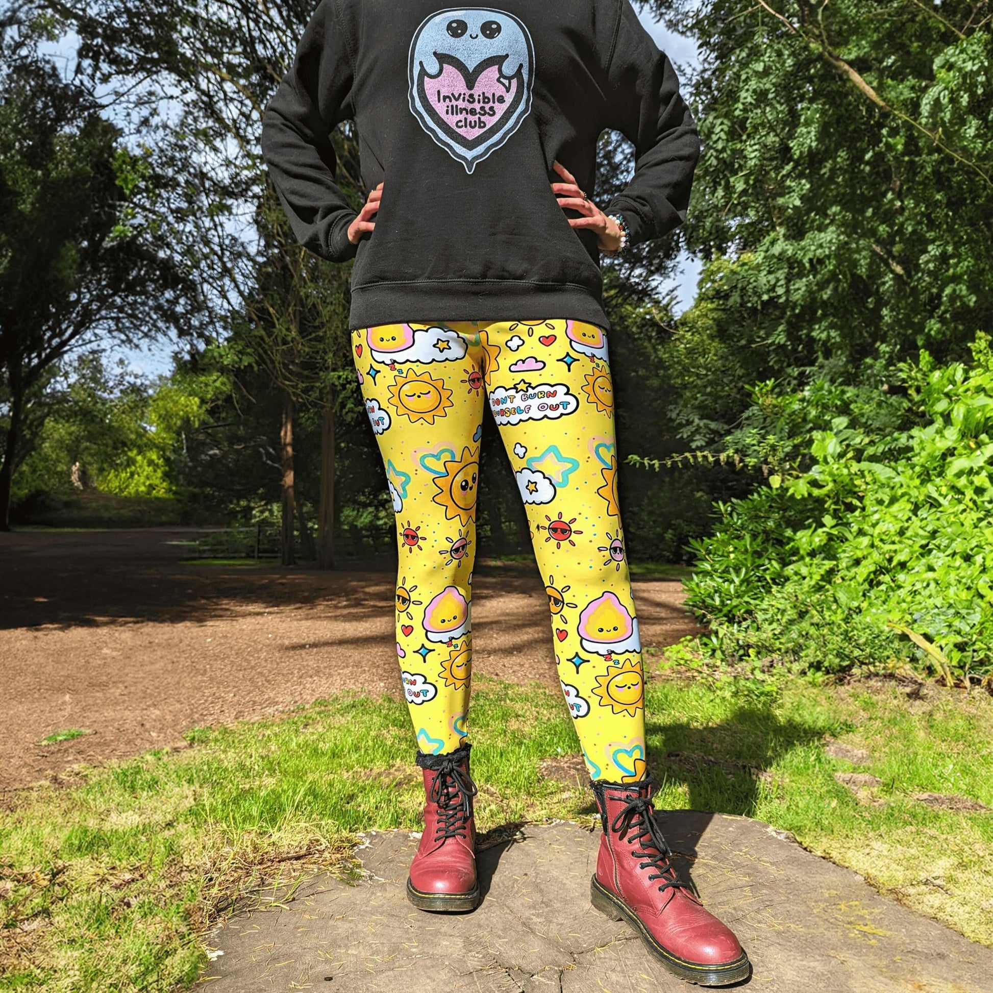 The Don't Burn Yourself Out Leggings being modelled by Nikky Box with an invisible illness club ghostie black sweater and red boots outside on a tree stump in the forest. She is facing forward with her hands on her hips, photo is cropped from the neck down. The print is yellow with various kawaii face sunshines, sparkles, hearts, pink flames and pink, blue and white clouds with don't burn yourself out inside in rainbow. Design inspired by the self care movement.
