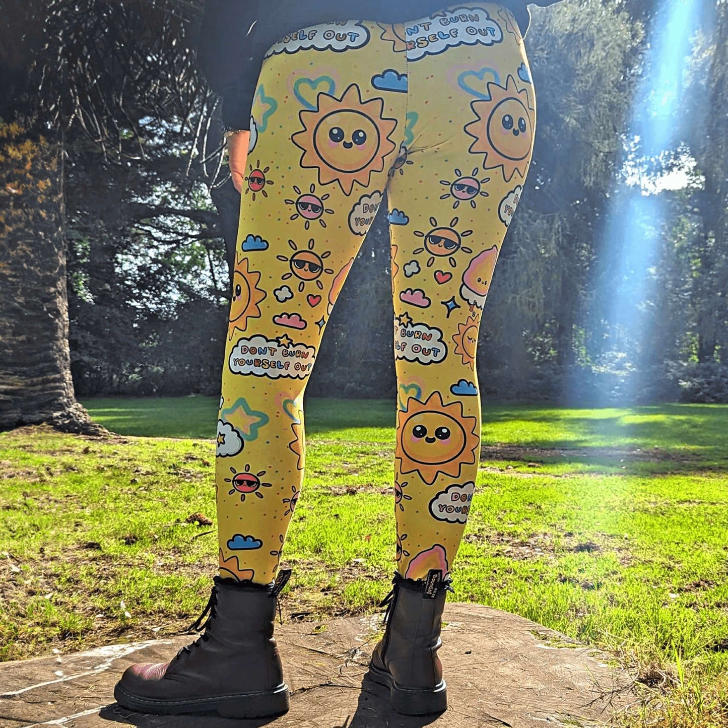 The Don't Burn Yourself Out Leggings being modelled by Nikky Box with red boots outside on a tree stump in the forest. She is facing away with her hands by her side, photo is cropped from the hips down. The print is yellow with various kawaii face sunshines, sparkles, hearts, pink flames and pink, blue and white clouds with don't burn yourself out inside in rainbow. Design inspired by the self care movement.