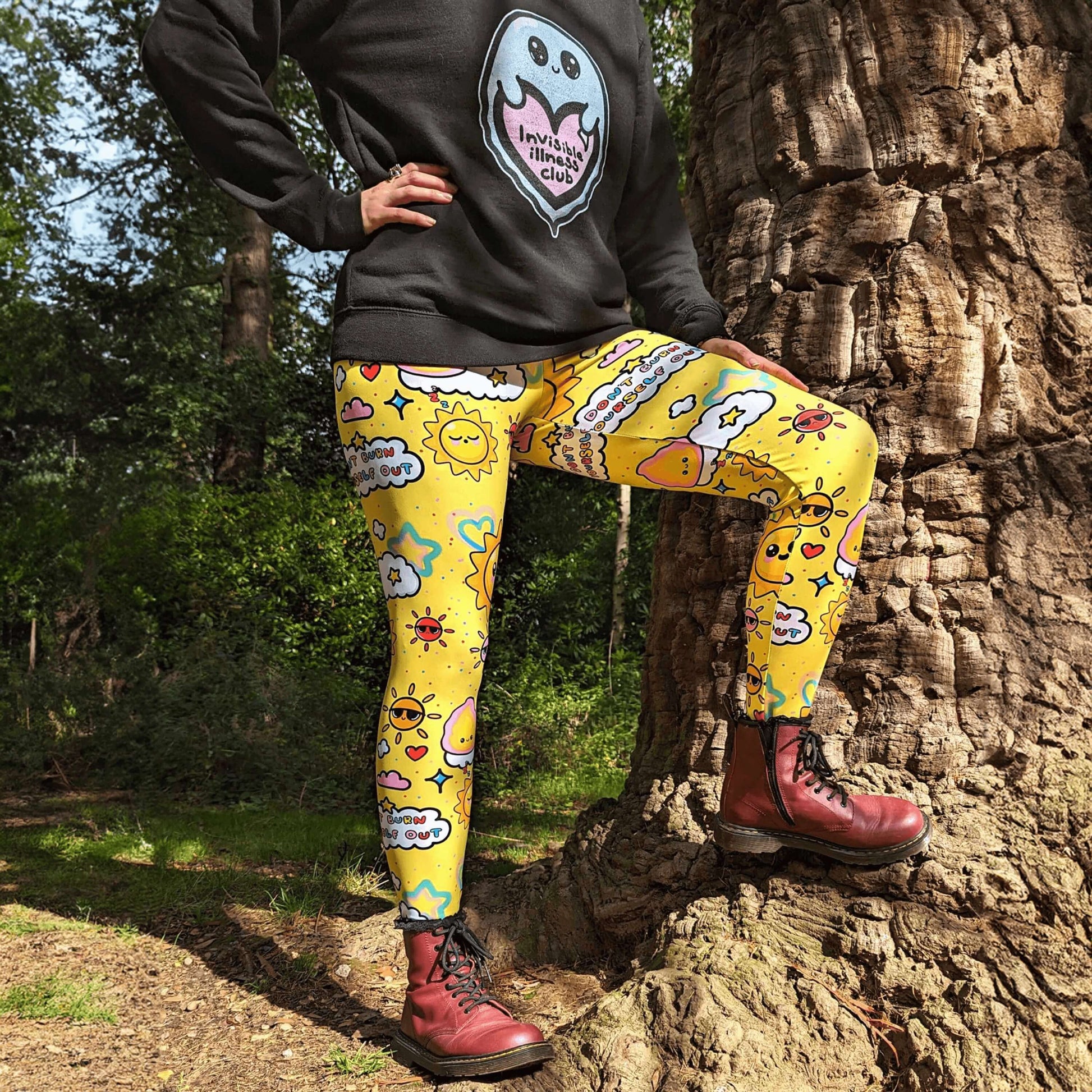 The Don't Burn Yourself Out Leggings being modelled by Nikky Box with an invisible illness club ghostie black sweater and red boots outside next to a tree in a forest. She is facing forward with one leg resting up on the tree and one hand on her hip, photo is cropped from the neck down. The print is yellow with various kawaii face sunshines, sparkles, hearts, pink flames and pink, blue and white clouds with don't burn yourself out inside in rainbow. Design inspired by the self care movement.